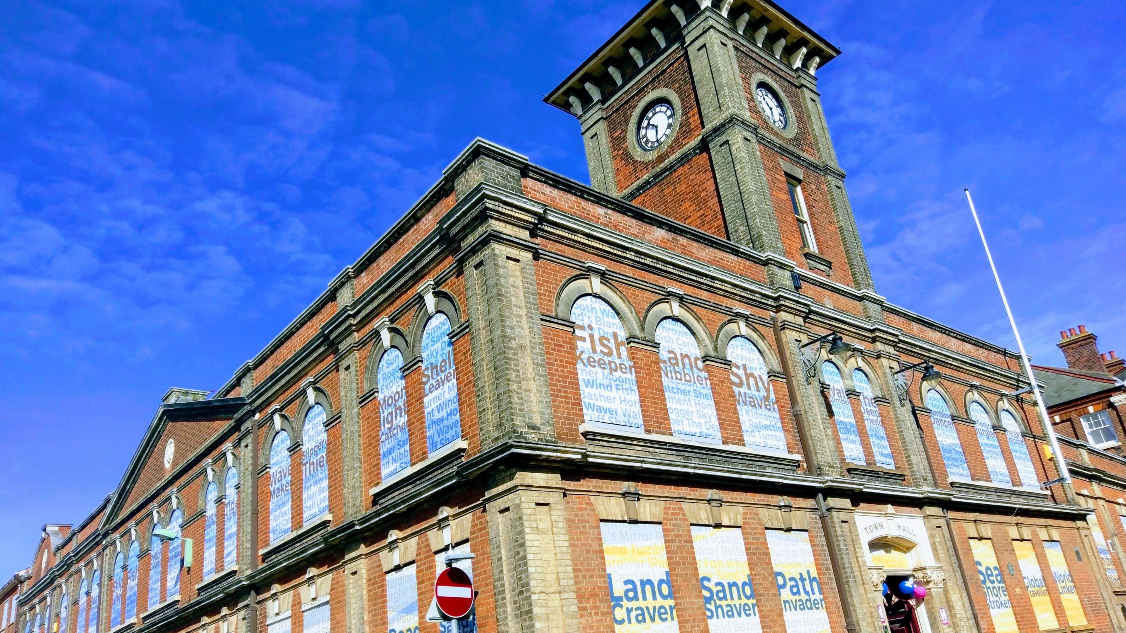Lowestoft Town Hall from the outside. It is a large building with a clock tower. All the windows have been covered with identical posters. On the ground floor these read 'Land Craver' and on the top they read 'Fish Keeper'.
