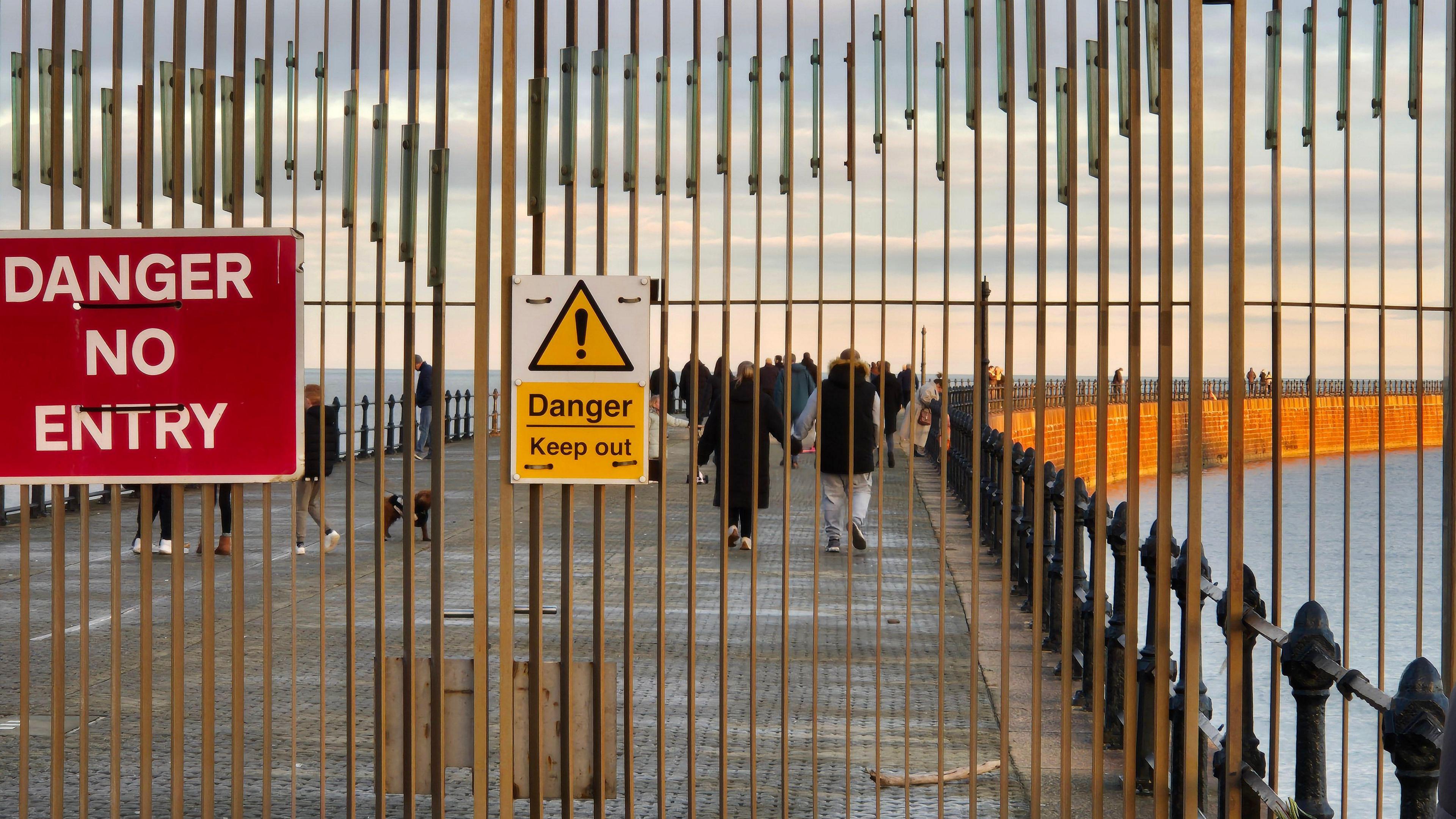 Roker Pier's gates with red sign reading Danger No Entry and yellow sign reading Danger Keep Out. Dozens of people are strolling along the pier behind the gates as the sun goes down. The sea is calm. 
