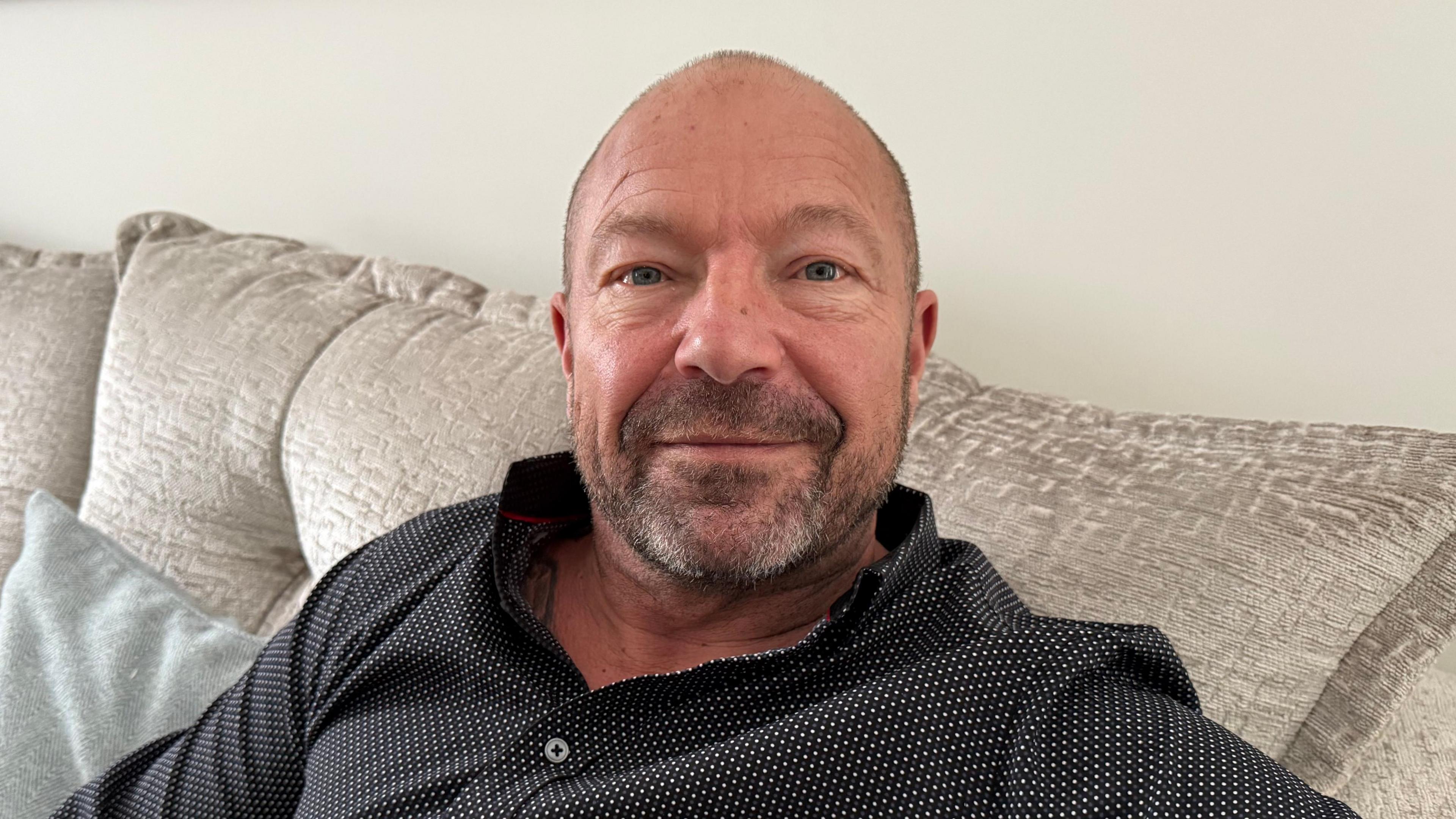 A tanned and bald man wearing a black and white polkda dot shirt sat down on a grey sofa. 