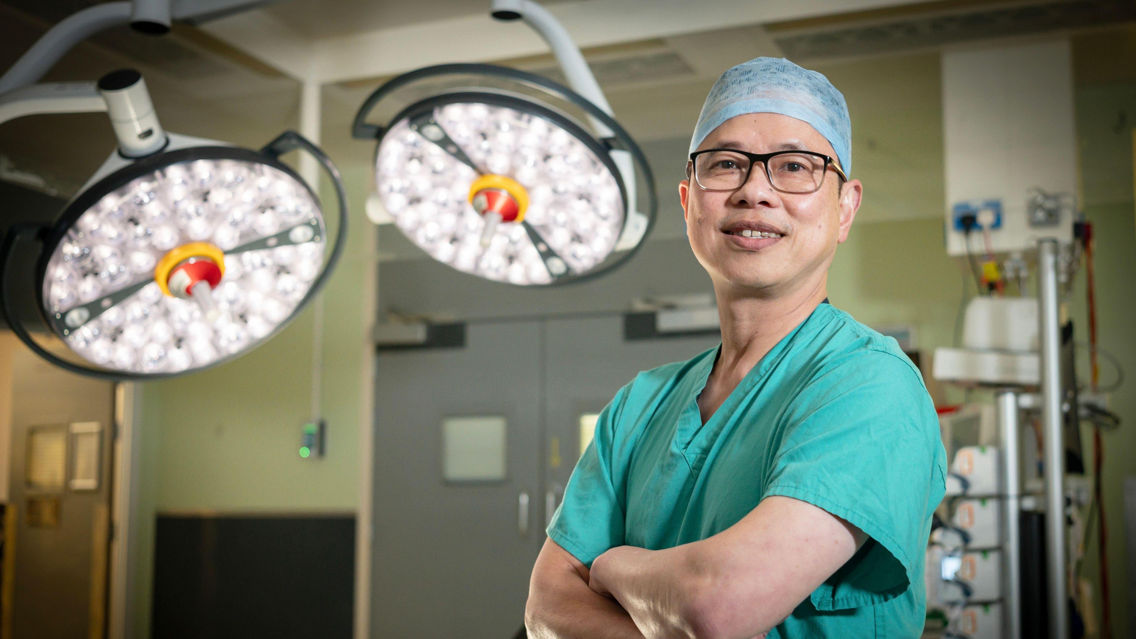 Dr Teik Choon See is standing in a modern operating theatre. He is dressed in surgical scrubs and a cap, and has black square framed glasses. Behind him is advanced surgical lighting and equipment. His arms are crossed and he is smiling at the camera.