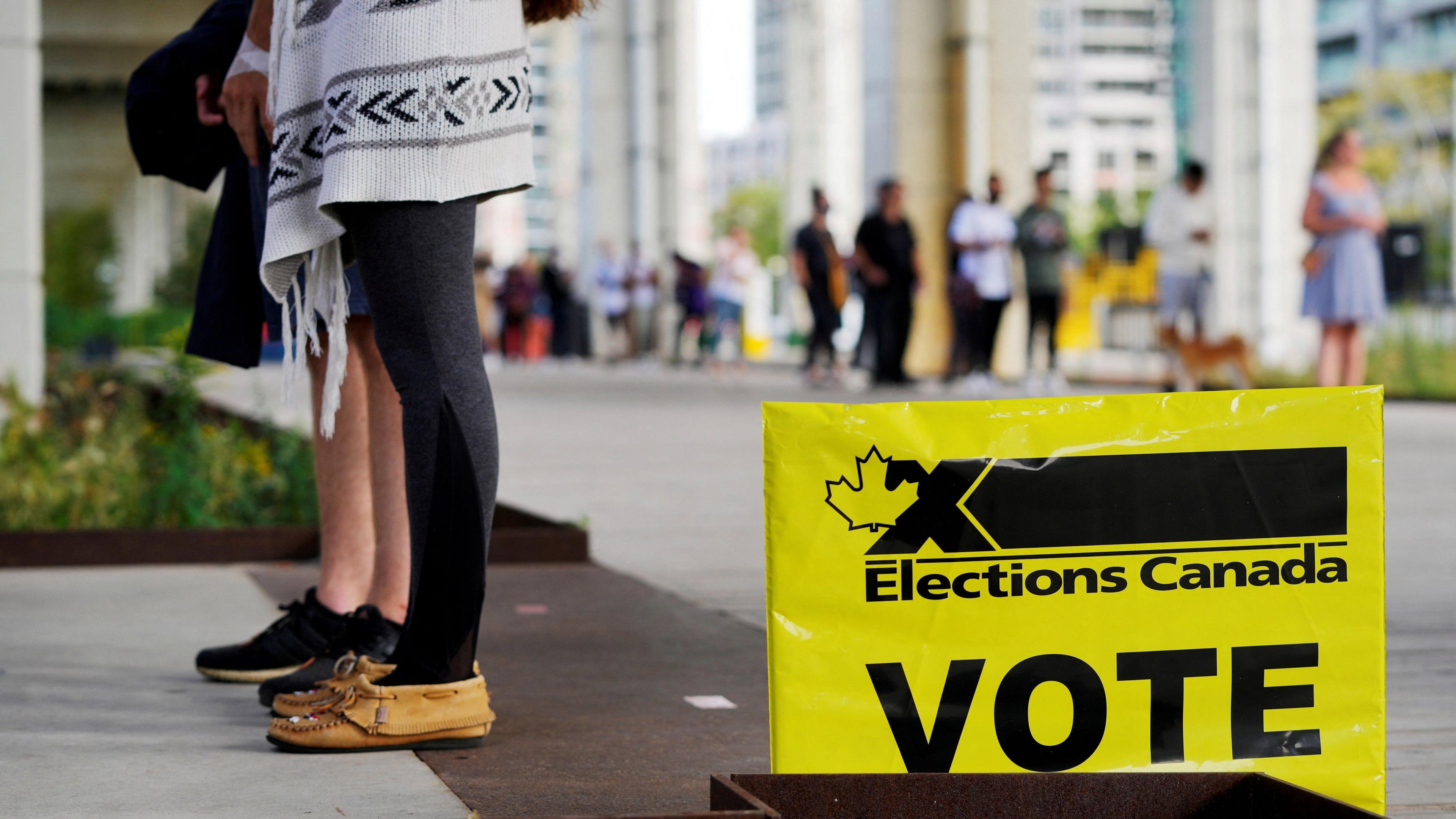 People are seen lining up near a voting centre. A bright yellow sign reads "elections Canada" and has the word "VOTE" in capital letters