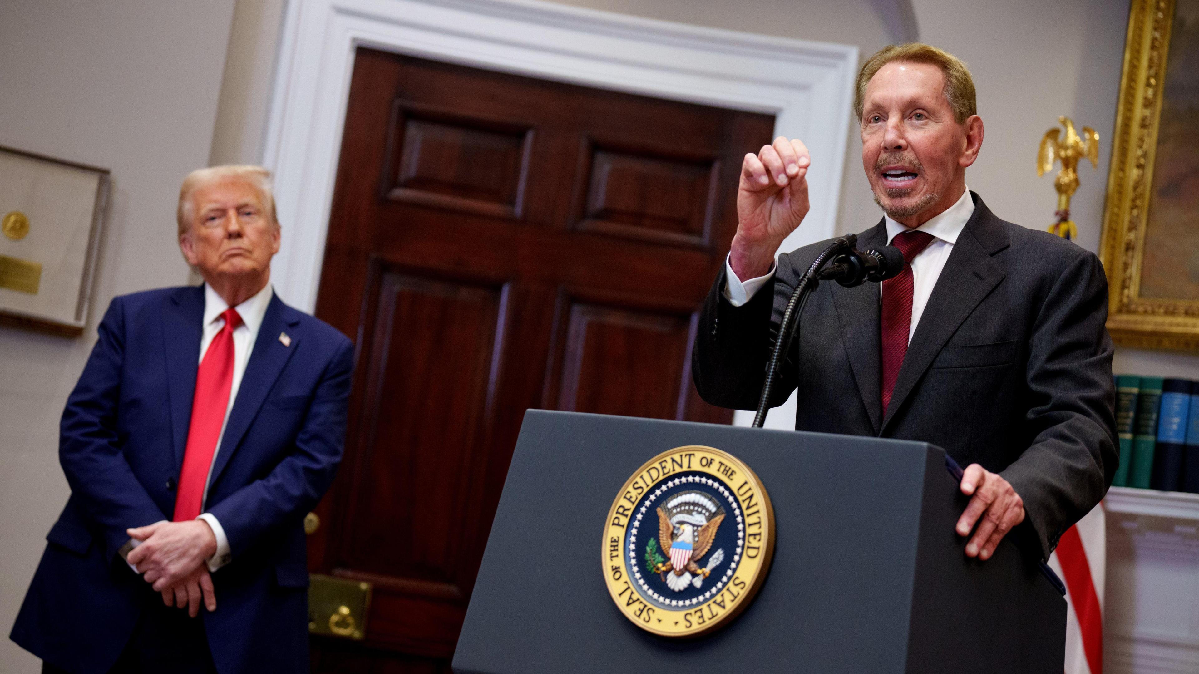 Larry Ellison at the White House with Donald Trump in January 2025. Ellison is at a podium in a dark suit with burgundy tie with one hand up for emphasis while Trump stands on the side, with his hands folded in front of him and his head cocked