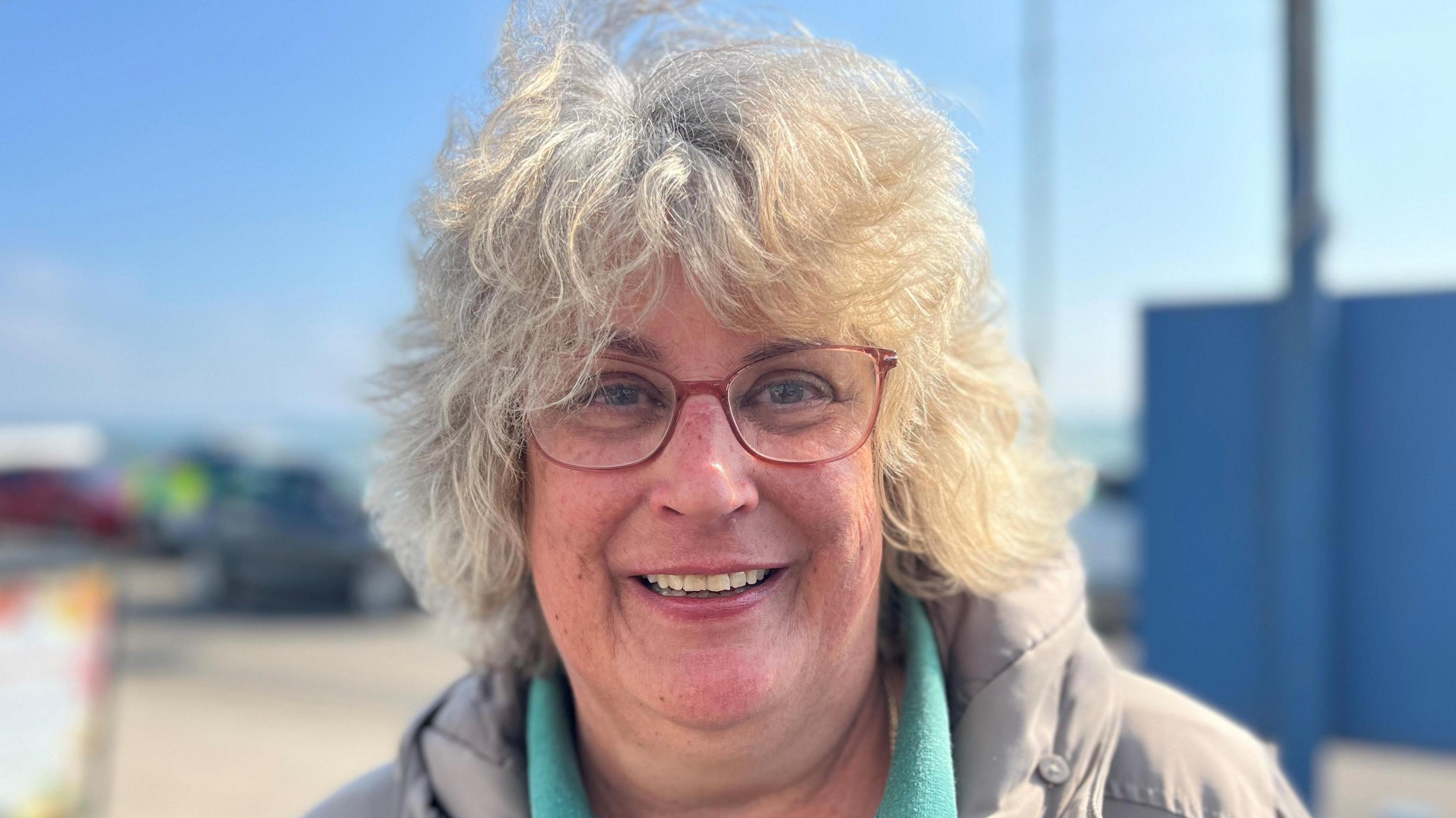 A lady looking straight at the camera with grey curly hair with a fringe, wearing red glasses a grey coat and green T-shirt, she is smiling and the sky is very blue.