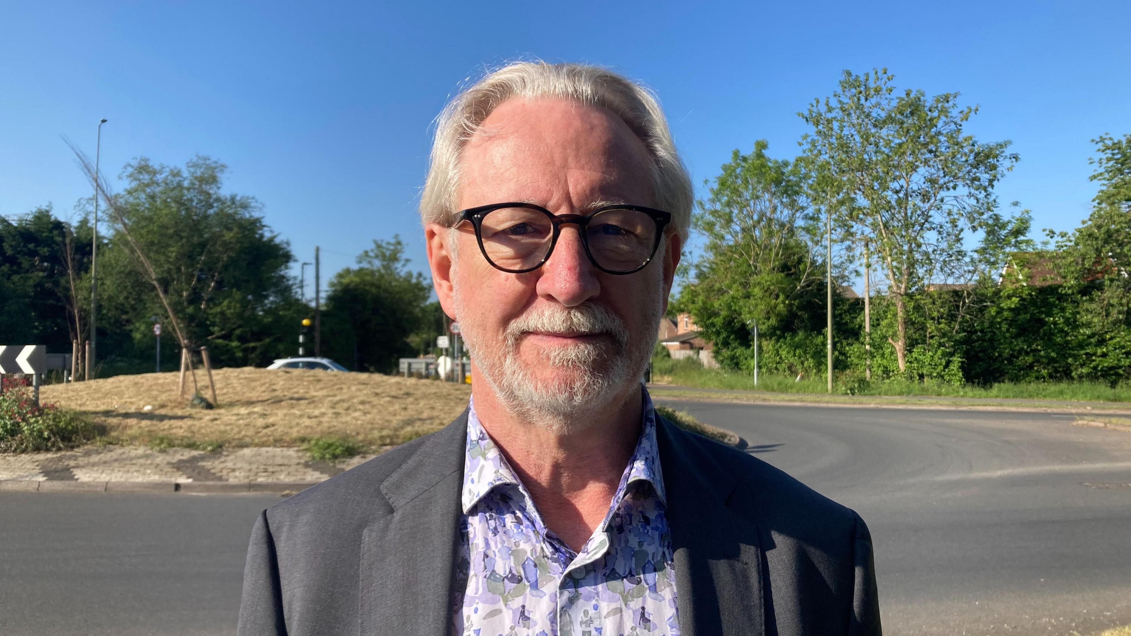 Martin Johnson poses for a photo. He is wearing a black suit jacket over a patterned shirt, with dark glasses. He has a neatly trimmed white beard and mid-length white hair. A roundabout can be seen in the background.