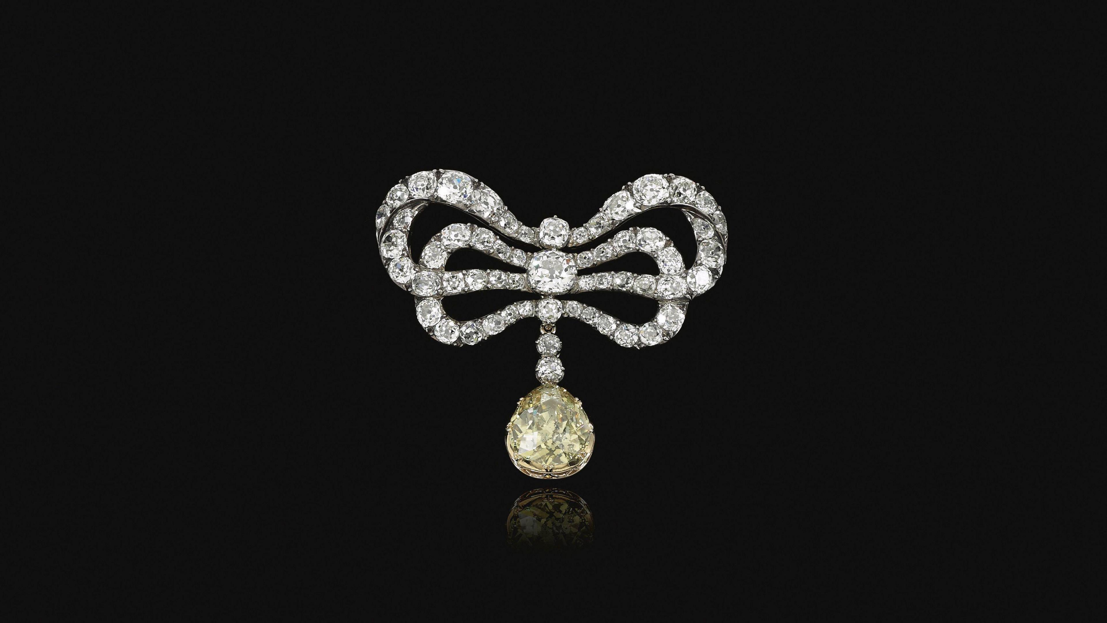 An ornate bow-shaped brooch made of diamonds, with a large yellow diamond drop pendant hanging from the centre, set against a black background.