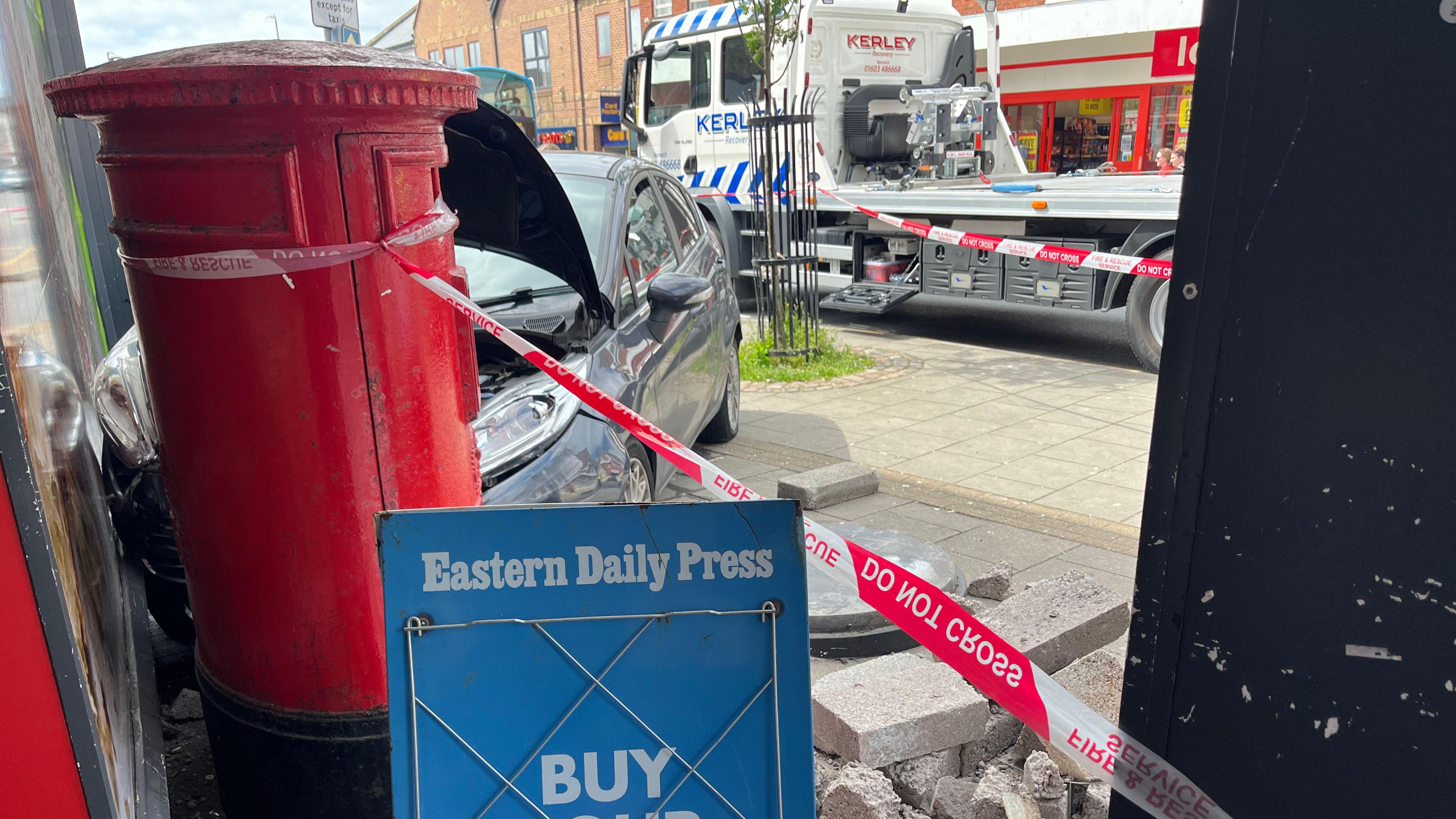 A car with its bonnet up, behind the red postbox, having crashed into a postbox