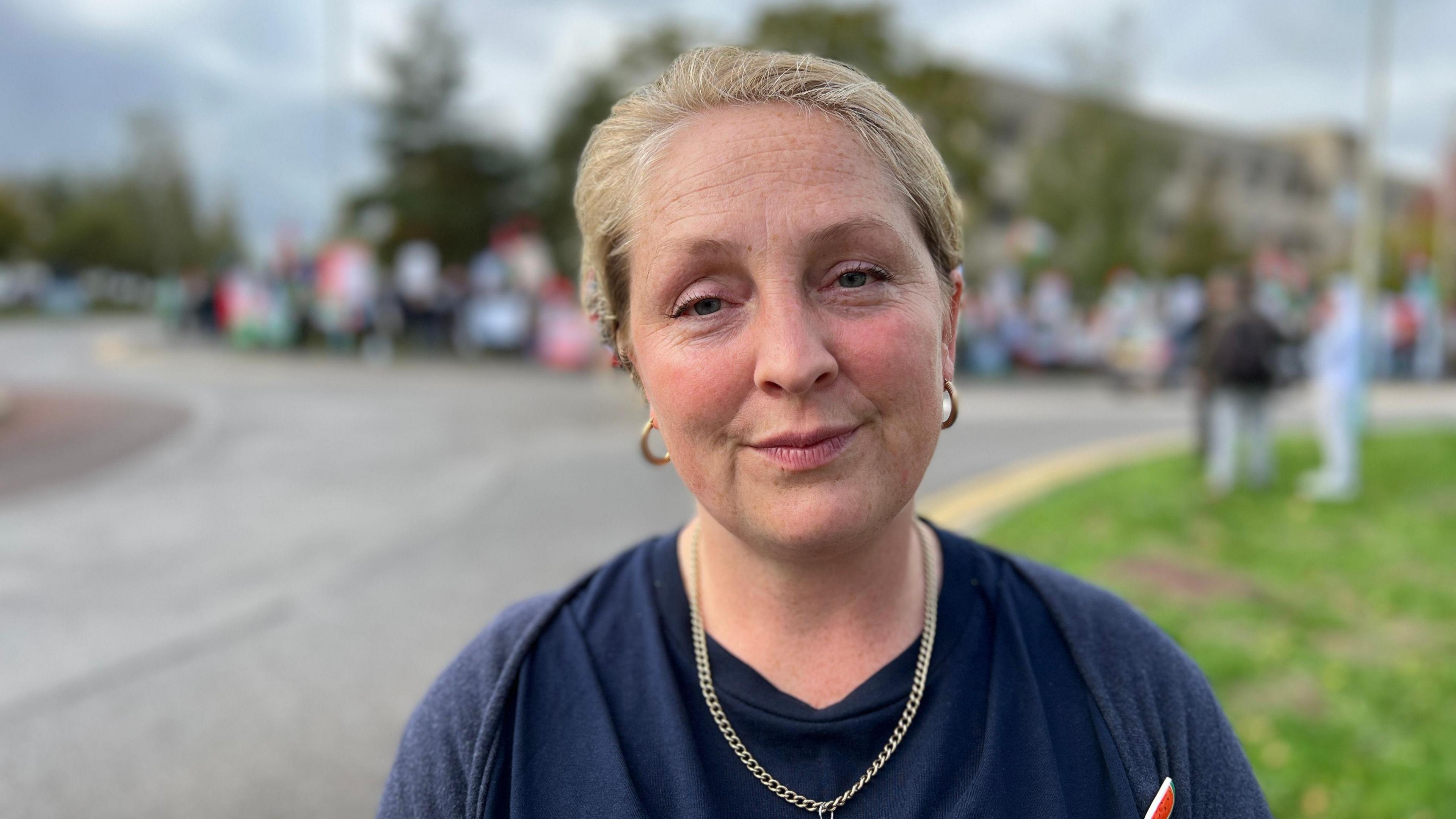 A middle aged woman with blonde hair tied back wearing a blue T-shirt, a gold chain and hoops, stands in front of an empty road. On the other side of the quiet road is a large, blurred group of protesters.