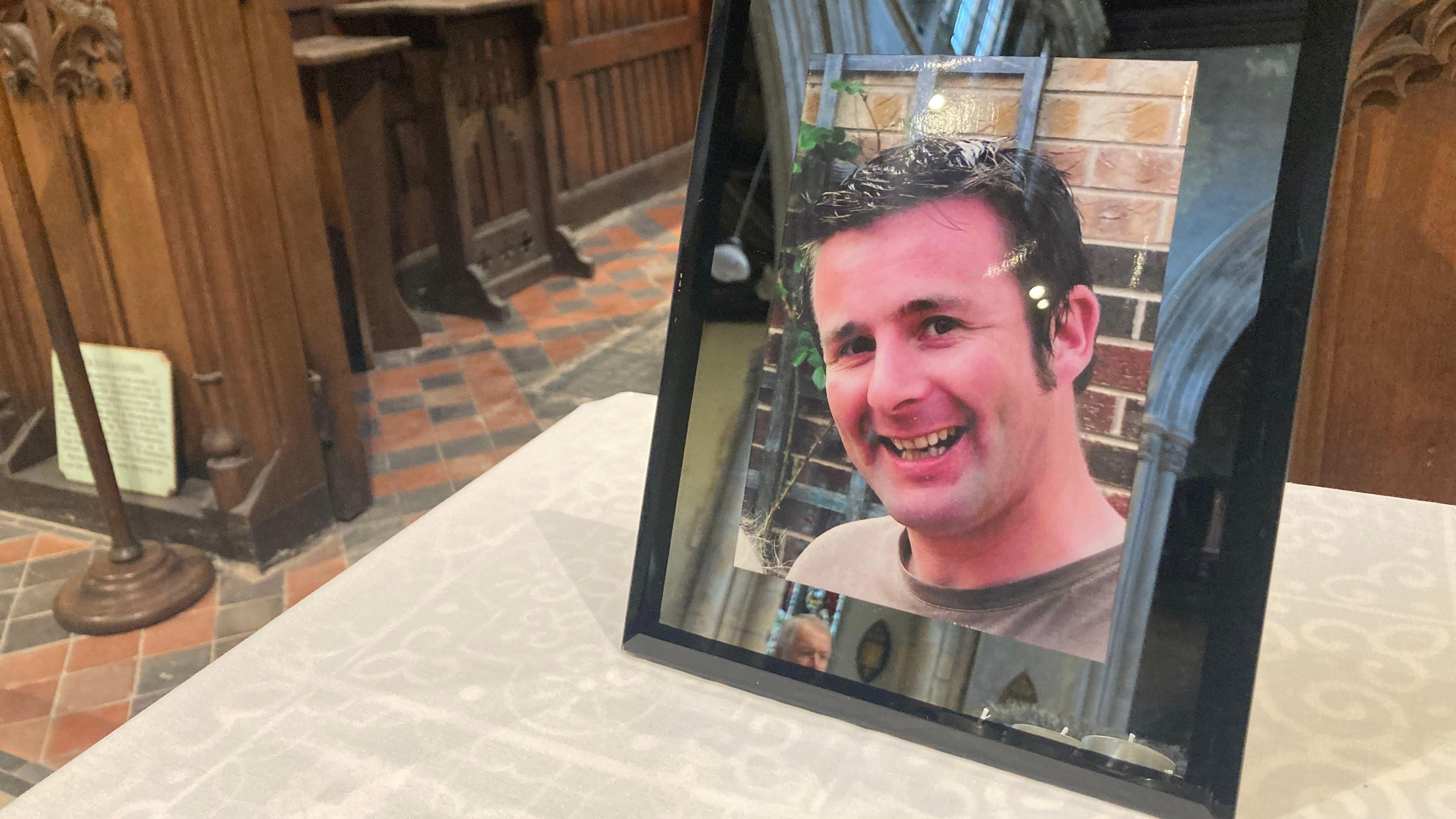A photo shows Danny smiling while looking into the camera. The frame has been put on a table with the pews in the background.