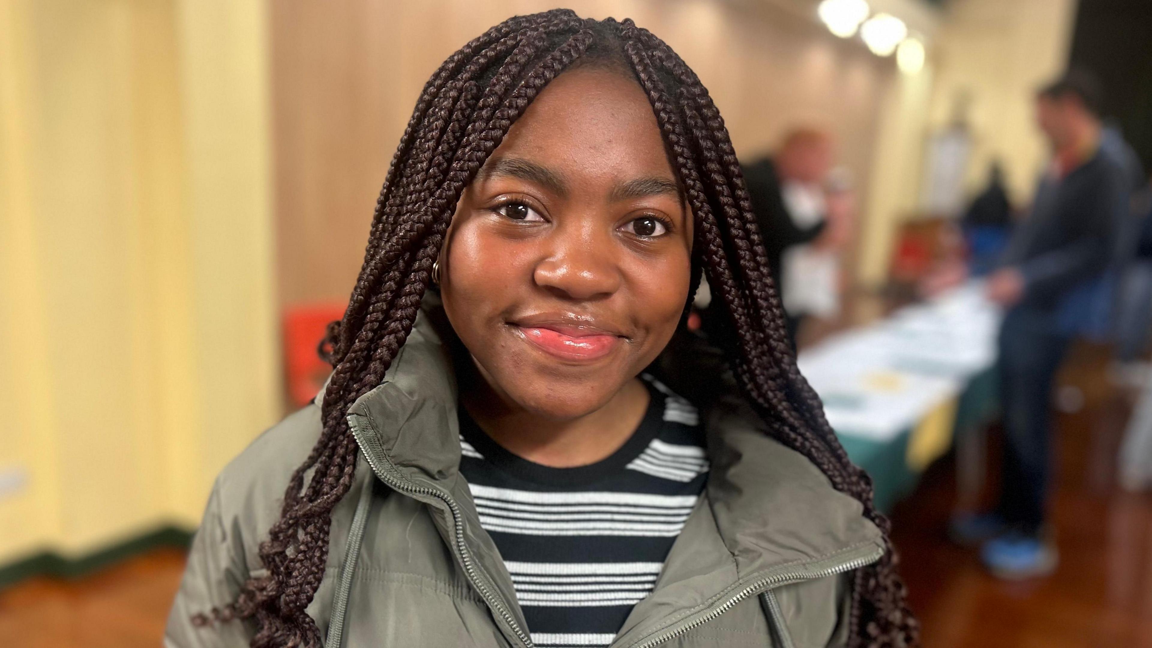 Lindi, a 16-year-old girl looking directly at the camera and smiling wering a grey coat and with a long table in the background.