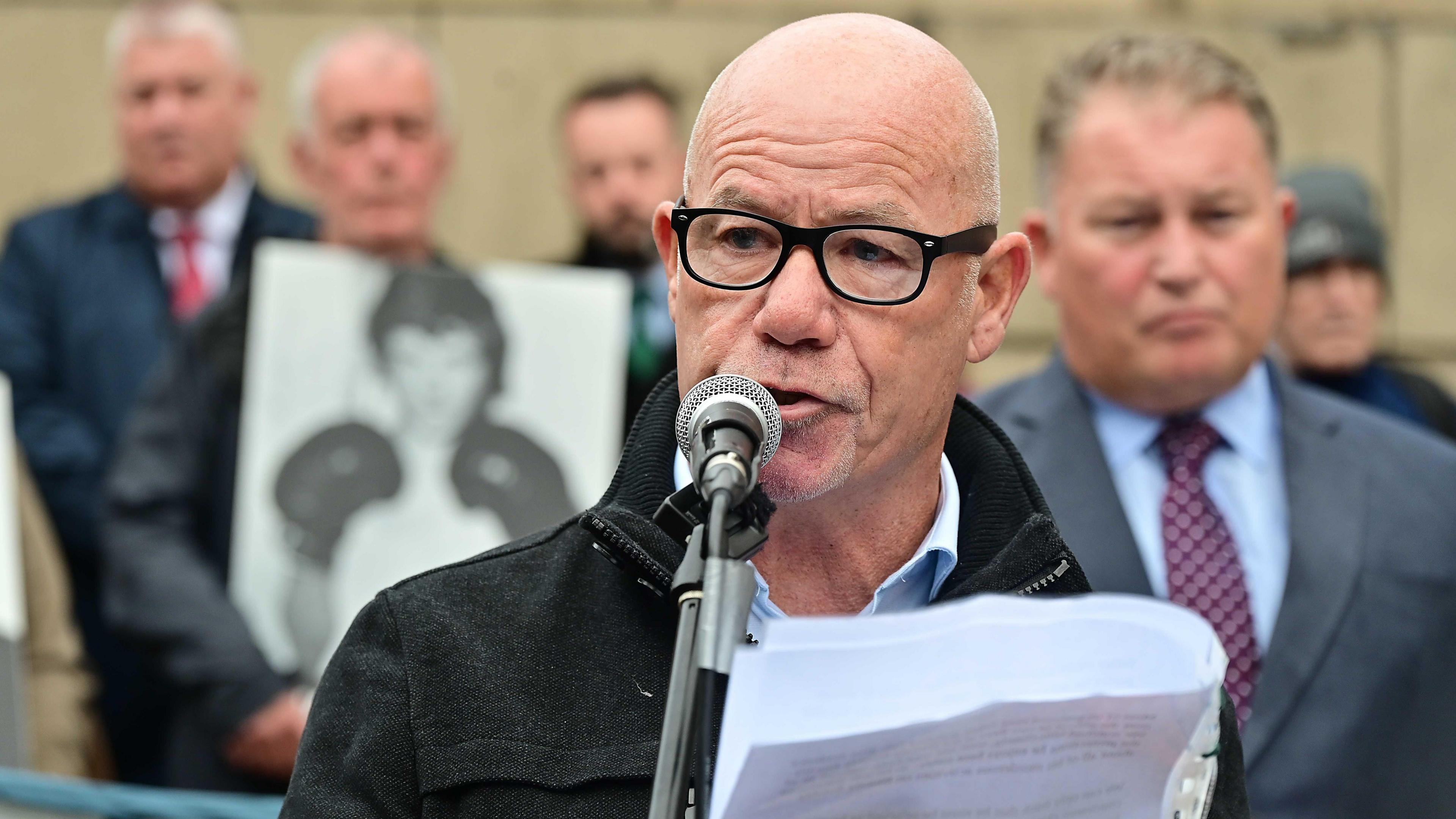 John McKinney wears a black jacket, blue shirt and black glasses. He is holding a piece of paper and standing at a microphone. There are people behind him, holding placards.