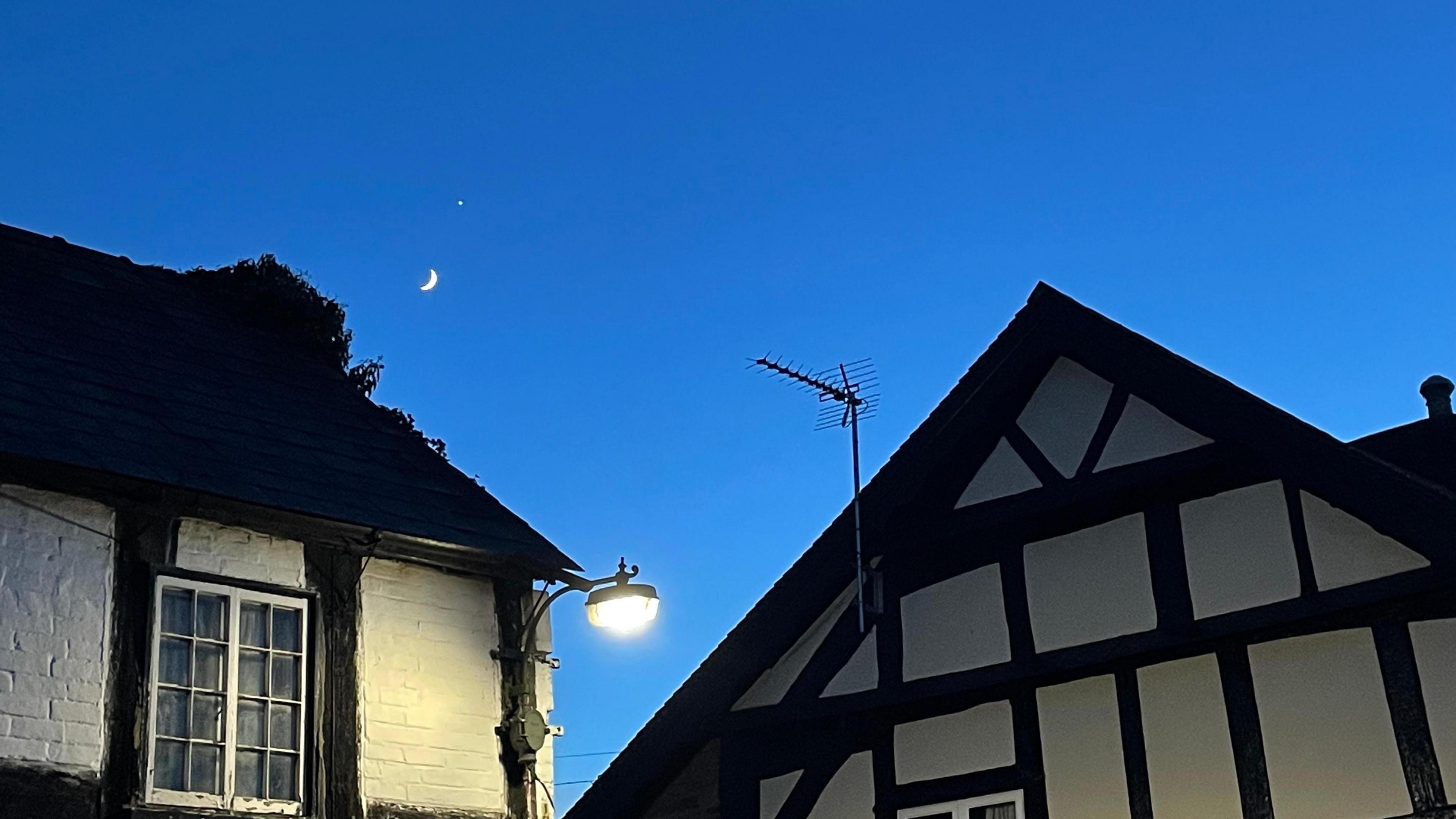 Venus and the Moon can be seen in a blue twilight sky over traditional black and white gabled houses. A street light shines from the roof of one of the houses.