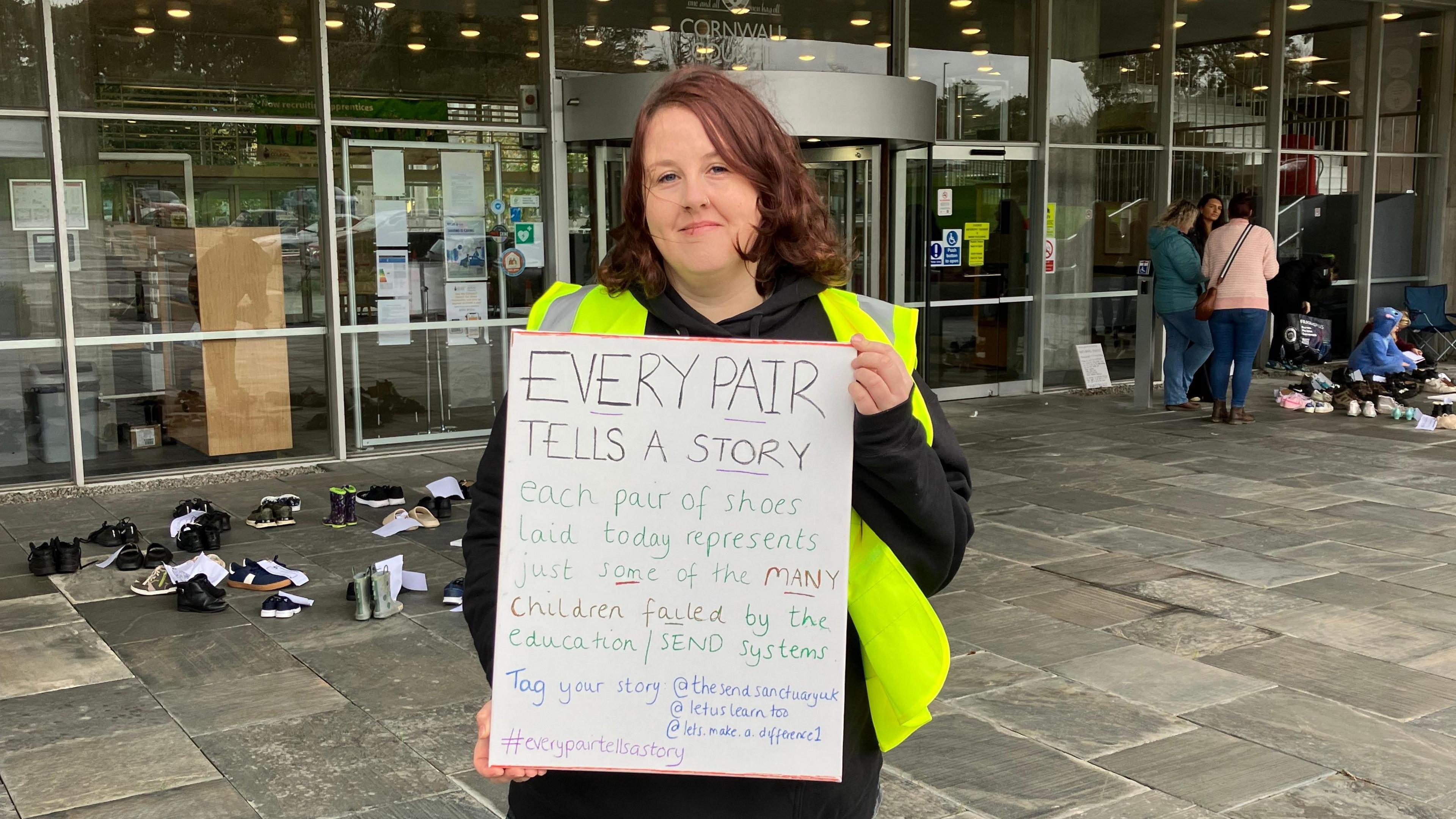 Kirsty Pellow is wearing a black hoodie, black trousers and a green hi-viz vest. She  has shoulder length brown hair. She is holding a whiteboard which says "EVERY PAIR TELLS A STORY." Behind her is County Hall, a building made of glass with a glass revolving door. On the grey concrete are children shoes, behind her to the left and right. There are people to the right of her in the background. 