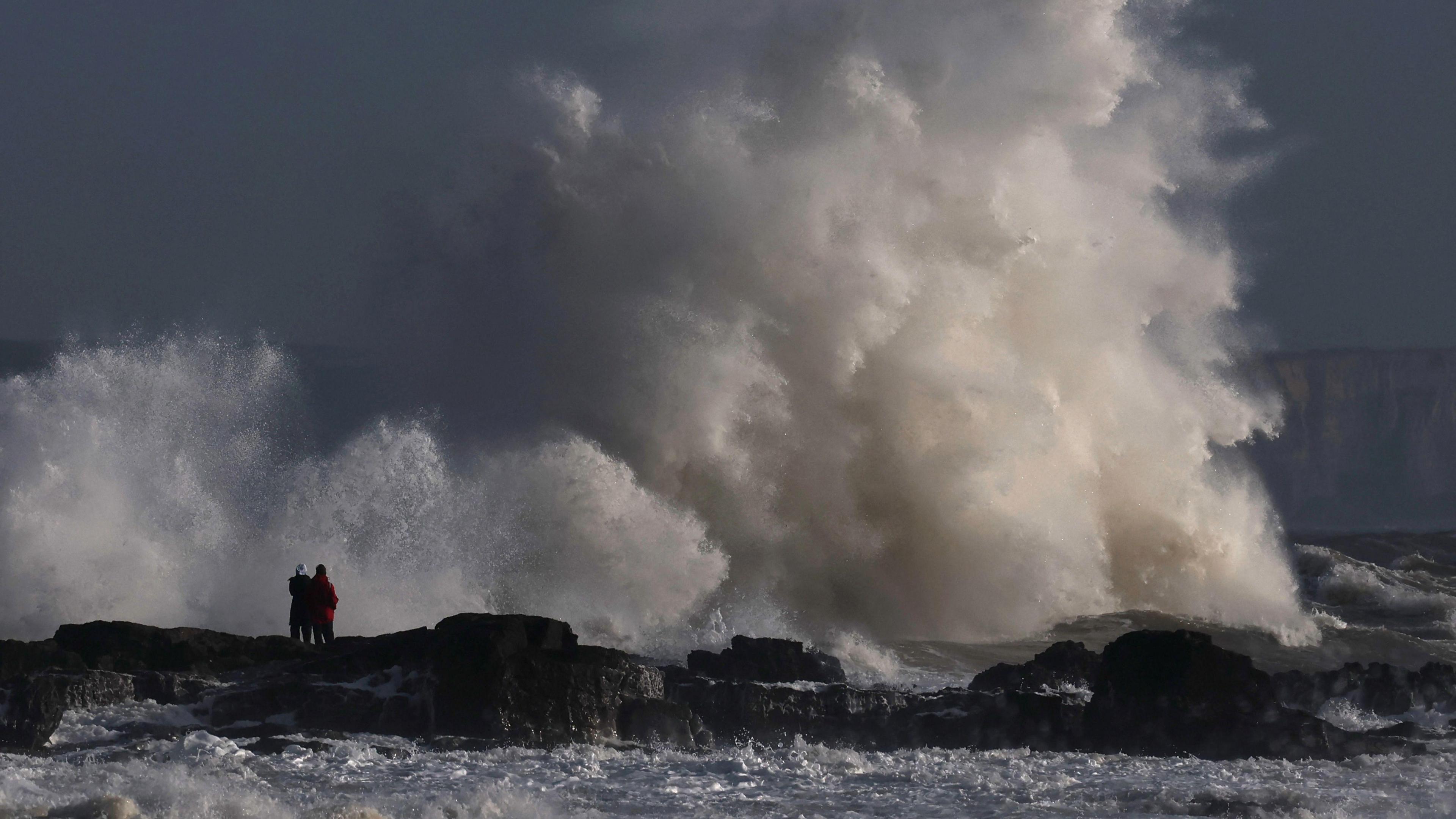 Large waves smash against rocks along the coast of Porthcawl, Wales, as two people look on