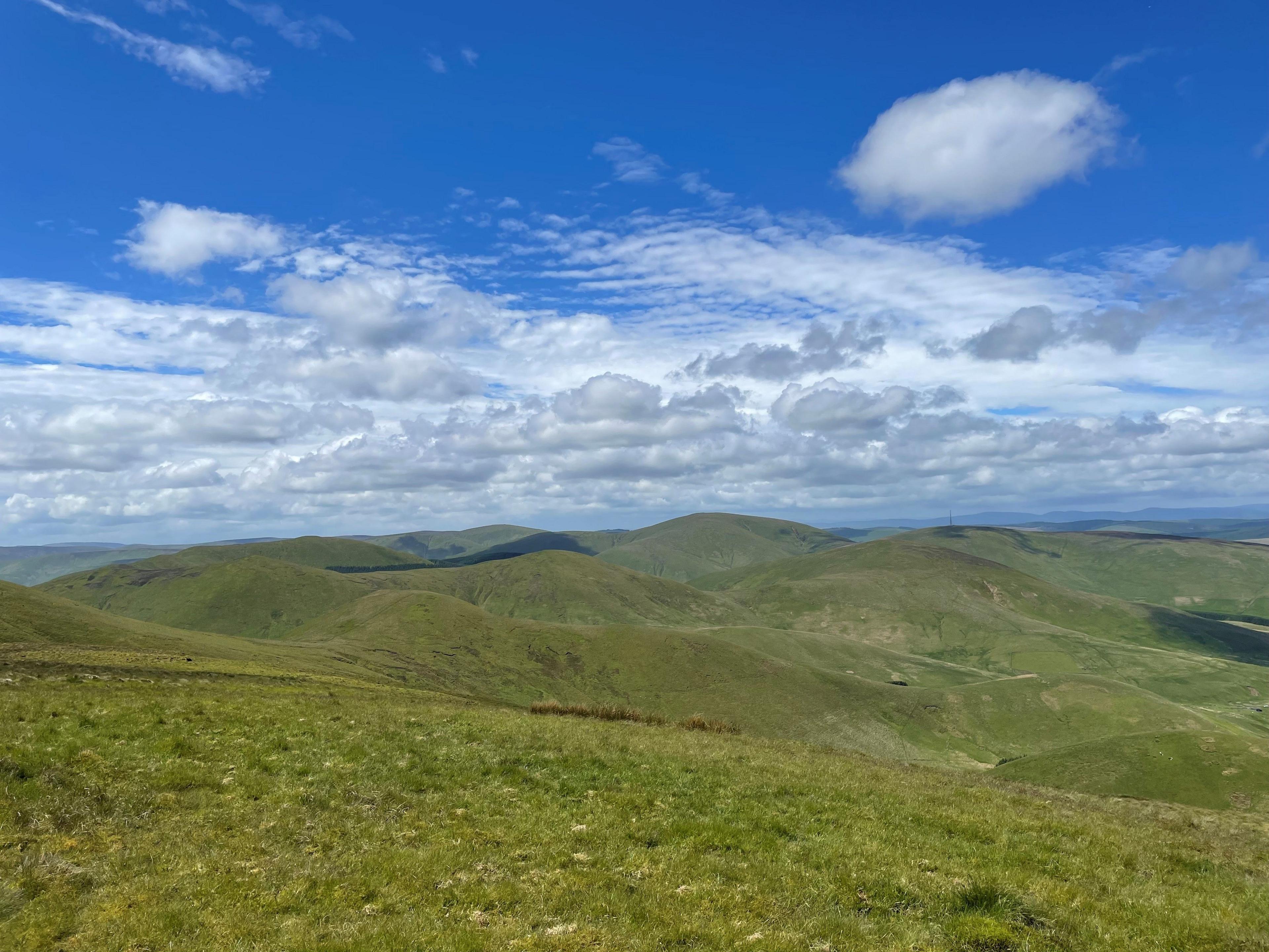 A set of rolling hills with low clouds and a blue sky above it where a wind farm could be built