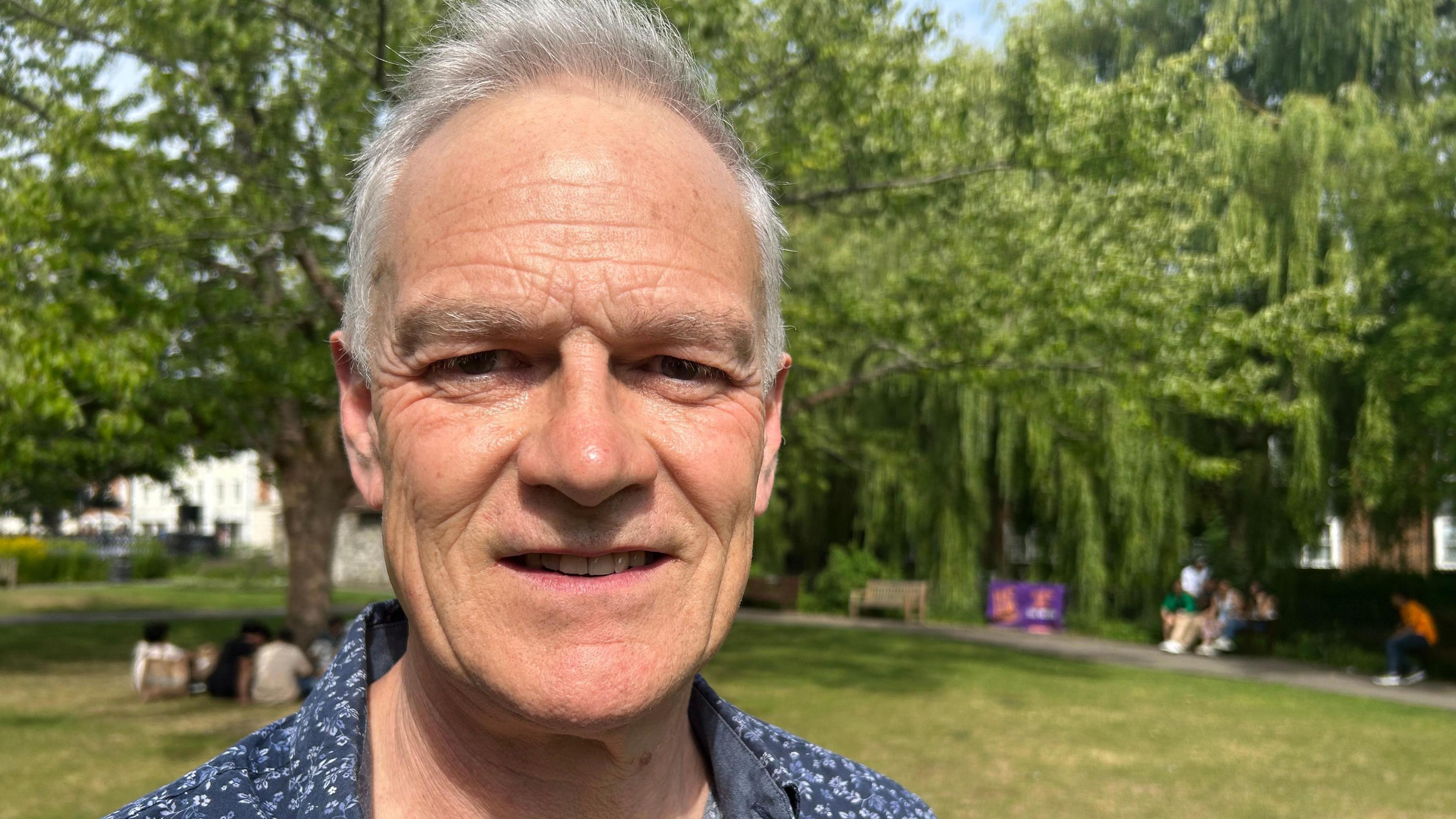 White man with white grey hair wearing a blue floral shirt looks at the camera. Behind him is a park with green lawn and a weeping willow tree'