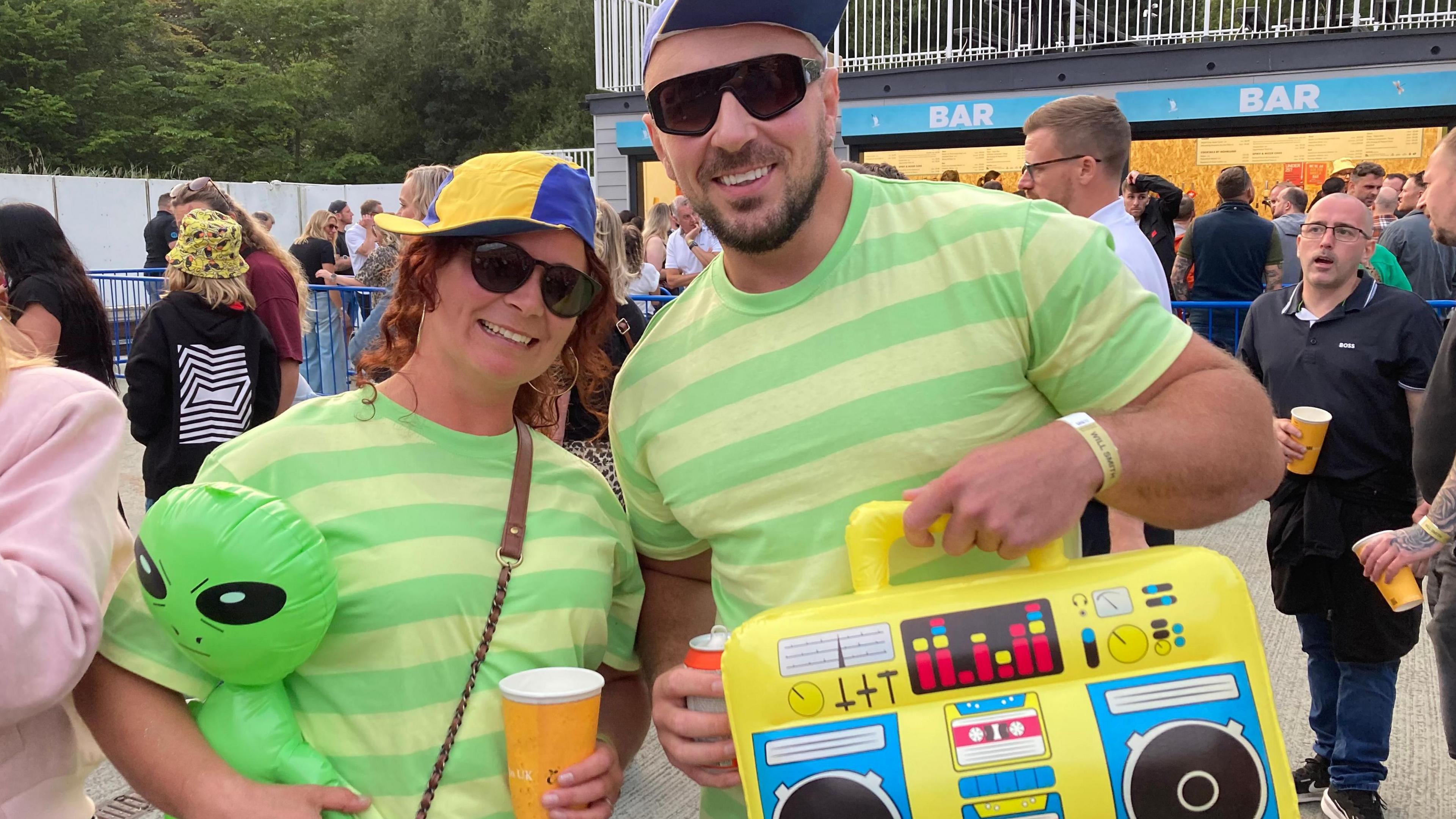 A woman and a man in green striped T-shirts. The woman is holding a blow-up green alien and the man is holding a blow-up boom box. There are other people milling around in the background.
