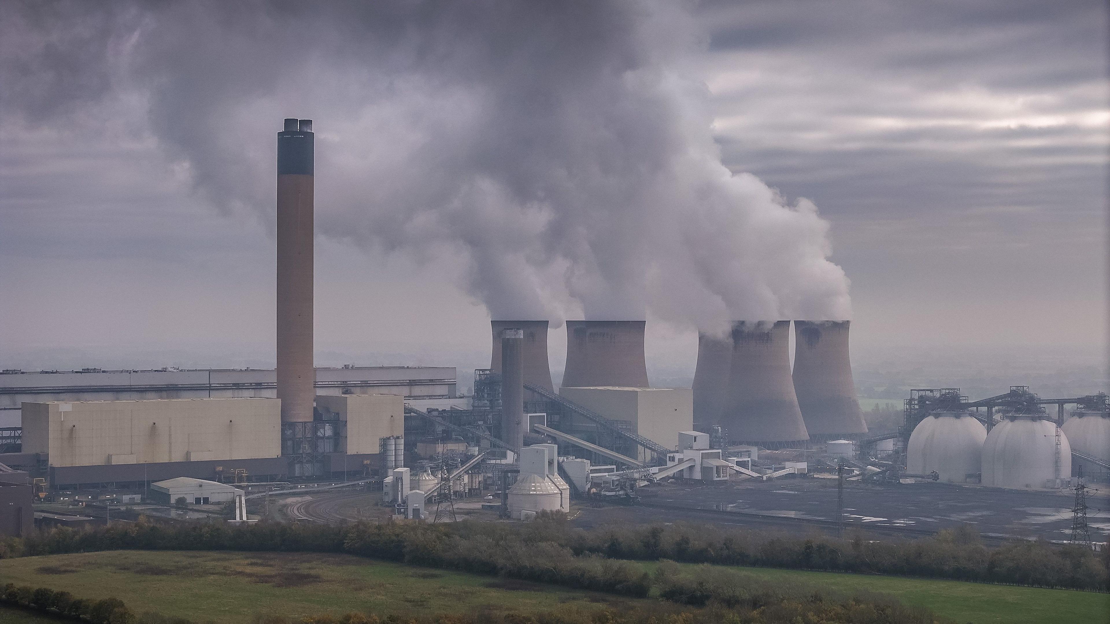 Drax power station. The photo shows smoke billowing out of six stout chimneys. The sky is cloudy and smoky. There are trees and grass in the foreground.