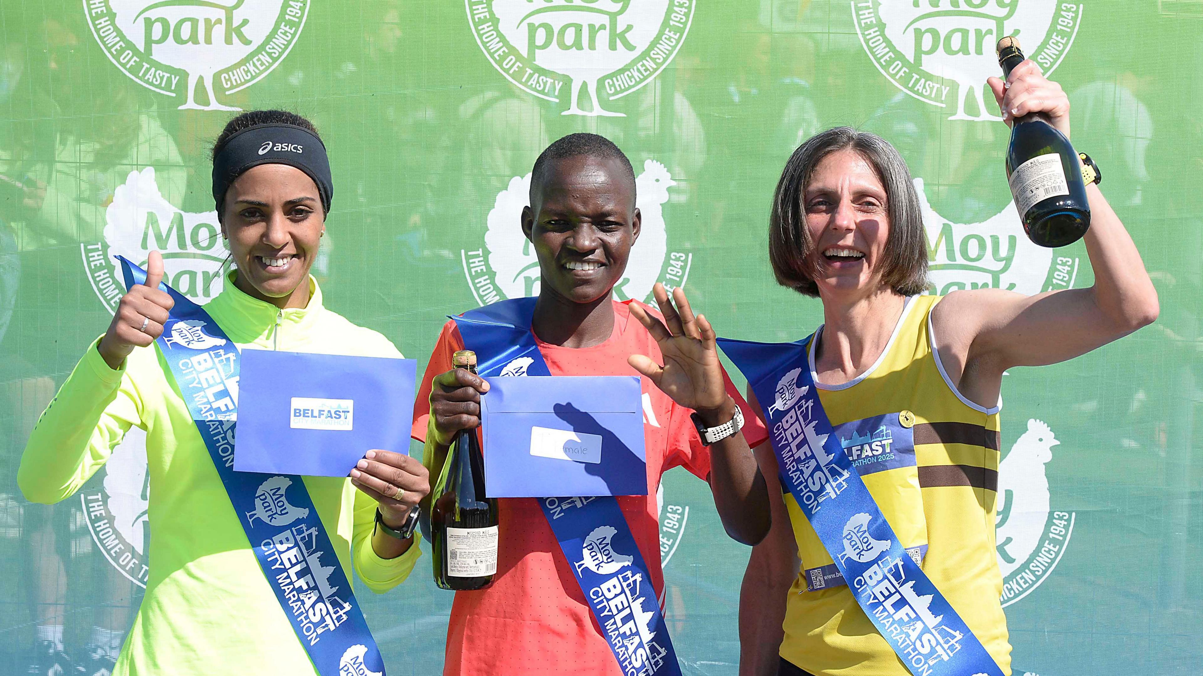 Three women stand in front of a green Moy Park screen at Ormeau Park after the Belfast Marathon. On the left Lalla Aziza wears a black Asics headband, a green zip up jacket and is holding a blue envelope, wearing a blue sash and is giving a thumbs up as she smiles at the camera. In the middle is Millicent Kibet, who is holding a bottle of champagne, a blue envelope and a blue sash. She is wearing a red athletic top and is smiling at the camera. On the right Gladys Ganiel is holding up a champagne bottle and is wearing a yellow athletic top. 