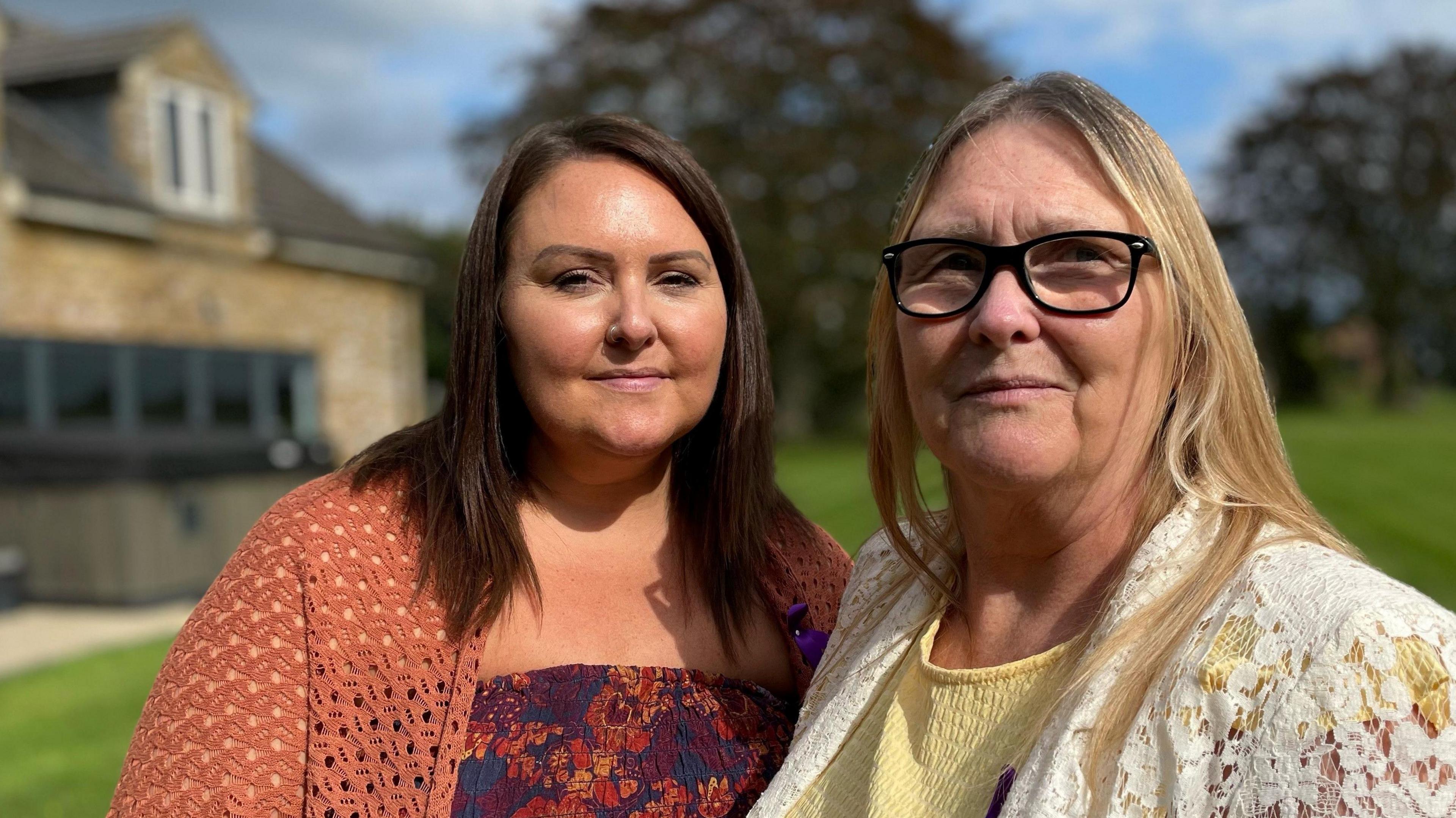 Two women standing in the garden of a house. Sarah-Jayne is wearing a dark orange knitted cardigan over a floral dress and has dark hair to just below the shoulders. Mum Annette is wearing a yellow dress with a white top. She has blonde hair and is wearing black-rimmed glasses.