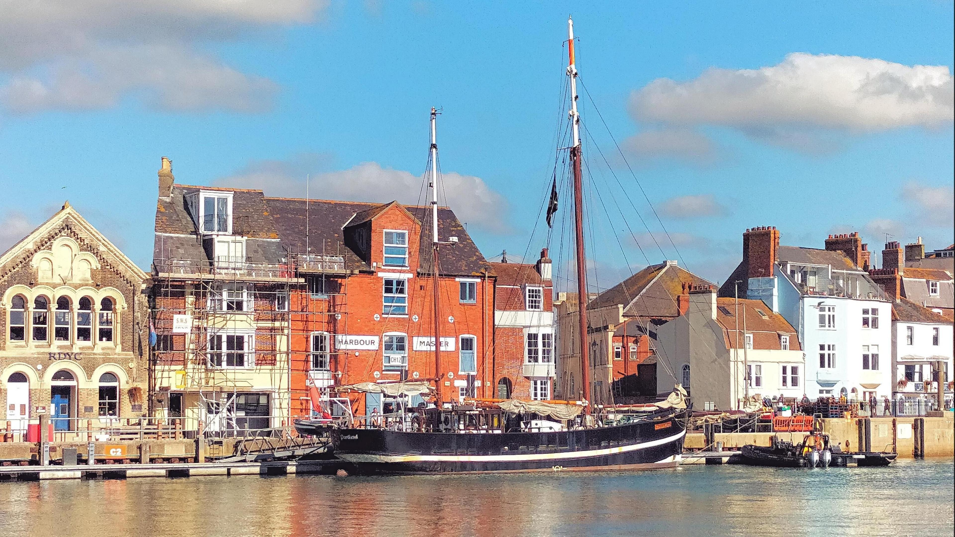 A black-bottomed boat with two masts is moored up in the harbour. Behind it sits a row of historical buildings - one has scaffolding on it. There is a bright blue sky with several white clouds. An inflatable dinghy with a motor can also be seen in the water, to the right of the boat