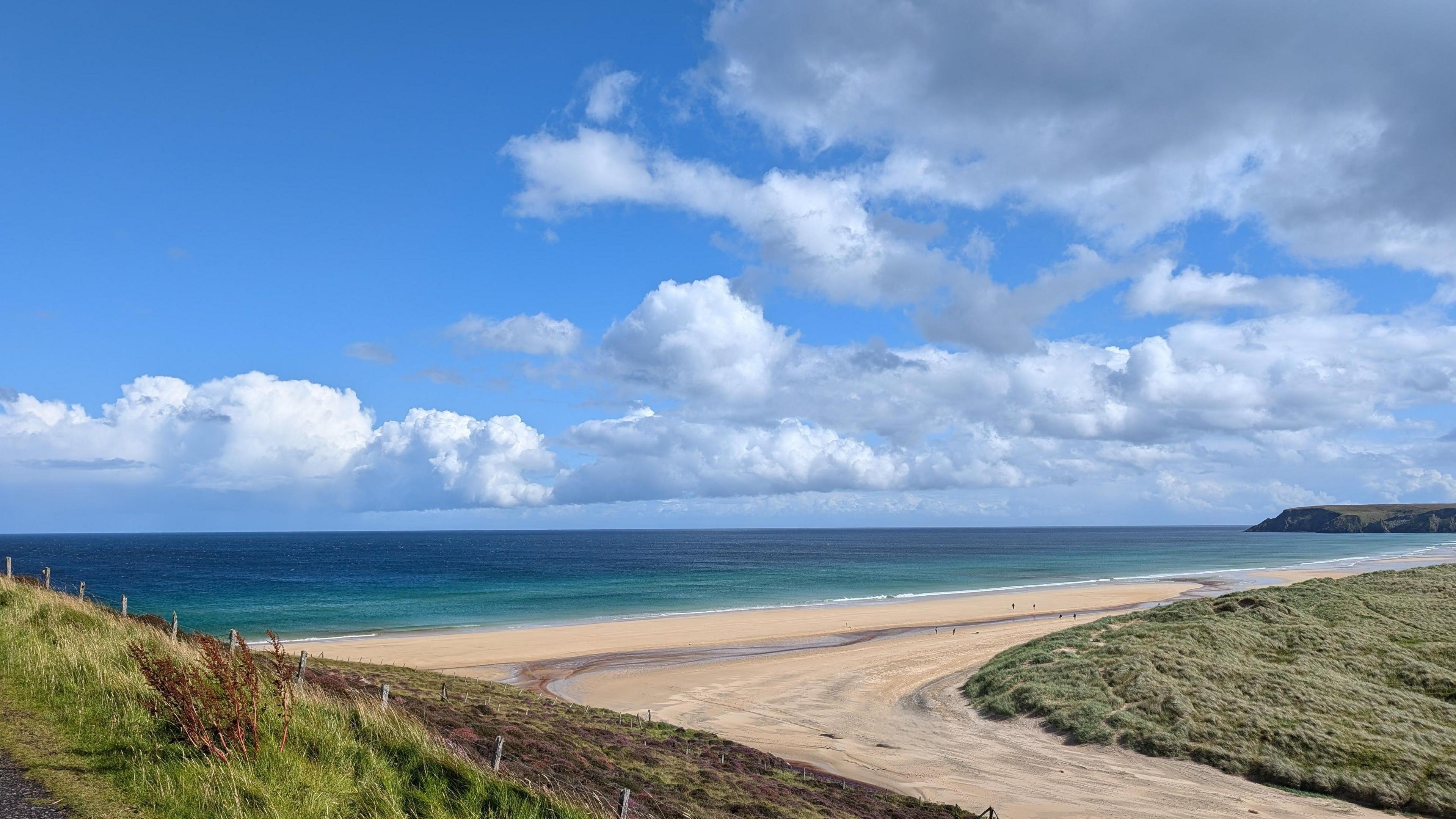 Garry Beach in the summer sun. The sky is full of white clouds and people can be made out as dots on the beach.