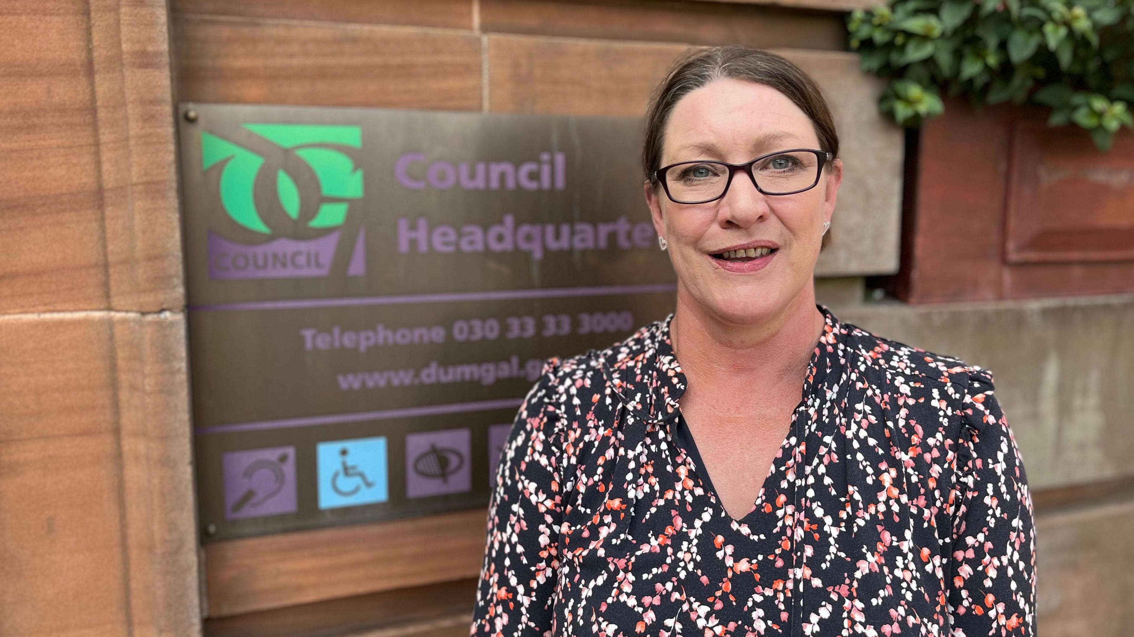 A woman with dark, pulled back hair and glasses in a patterned blouse stands in front of Dumfries and Galloway Council headquarters