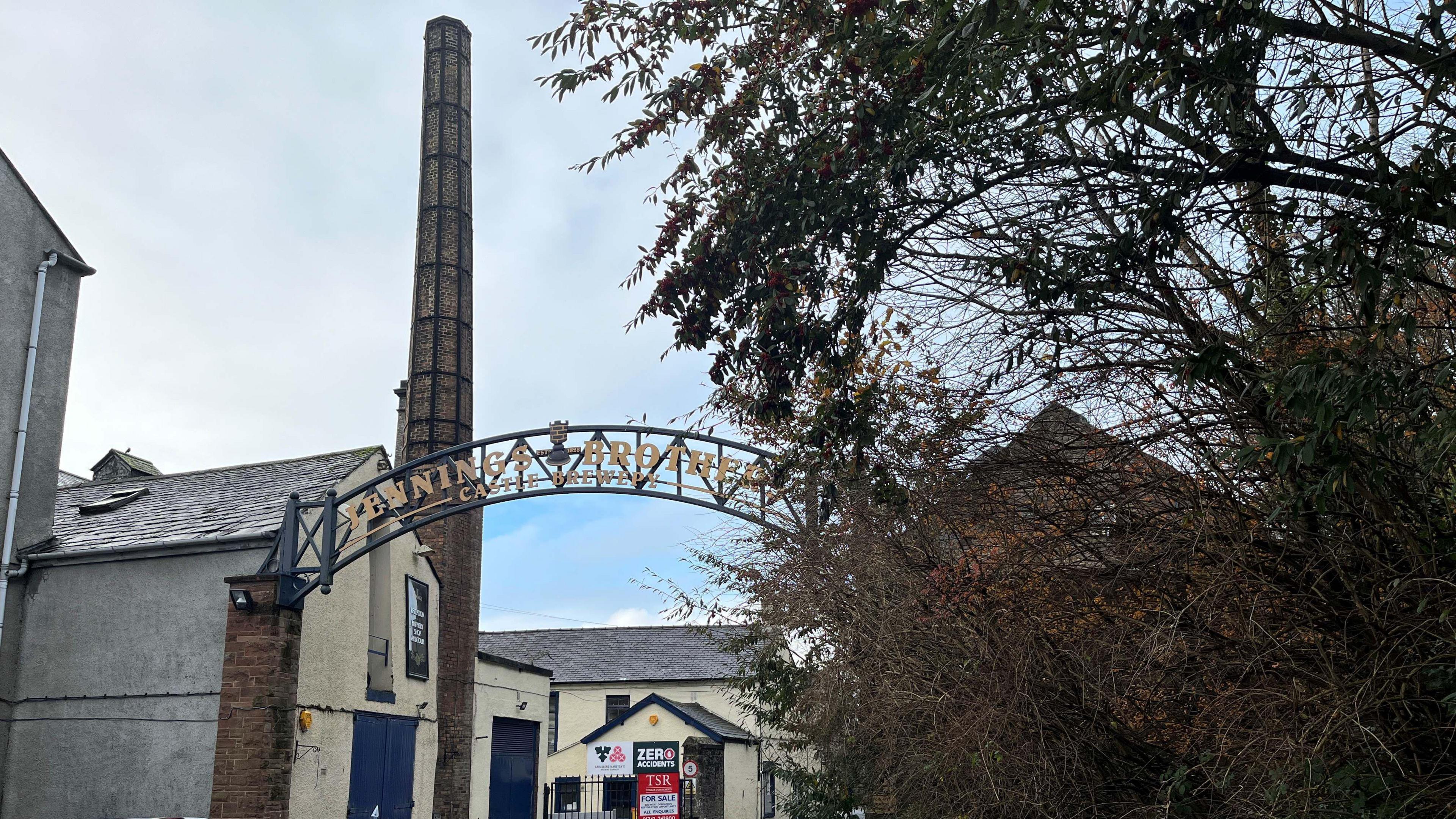 The entrance to the former Jennings Brewery in Cockermouth. There is a metal arch with golden lettering saying, Jennings Brothers, Castle Brewery.
There are industrial buildings and a tall chimney. A for sale sign can be seen on a gate.