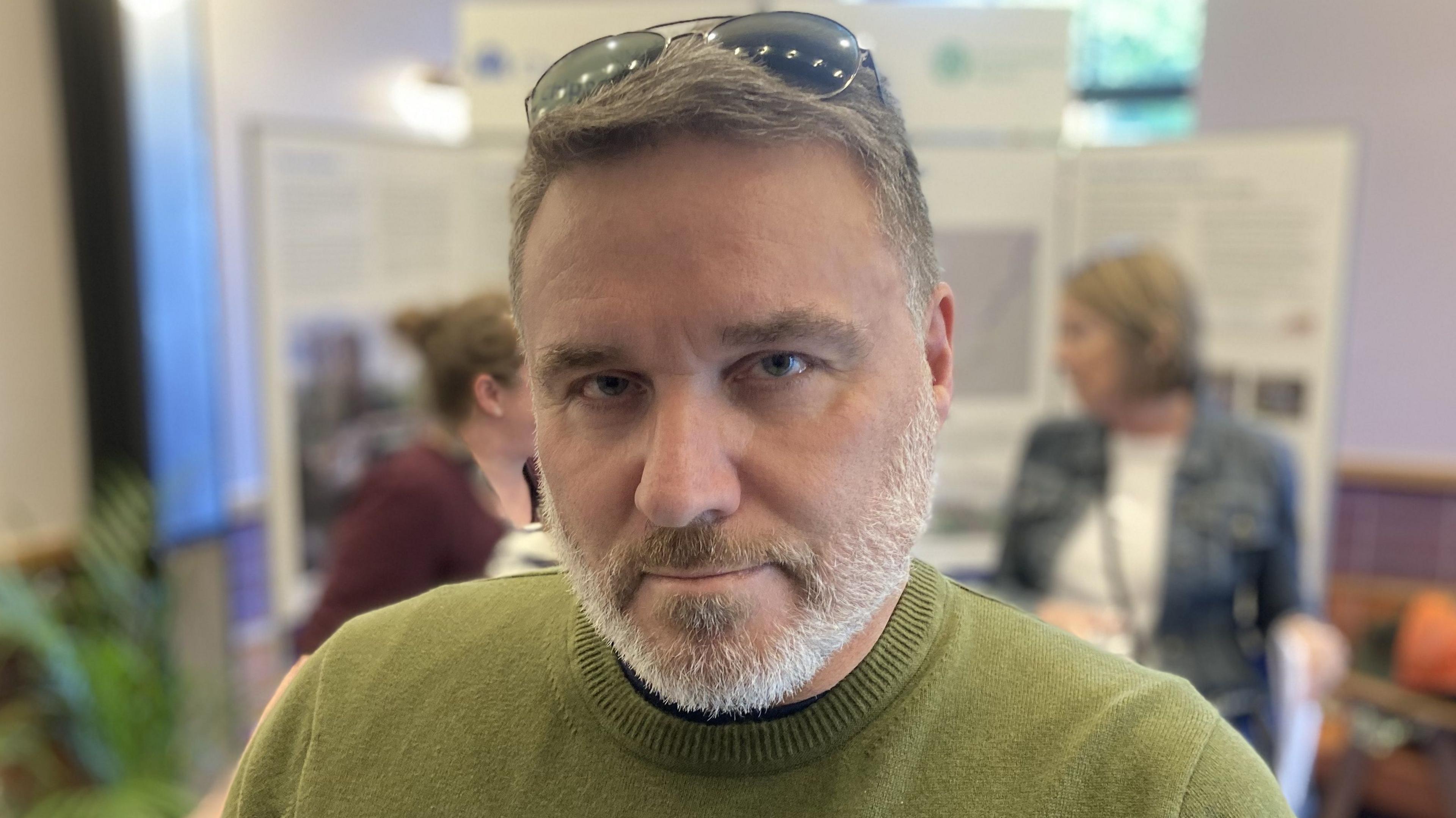 James Geoghegan looking stern with short grey hair, beard, wearing a green jumper