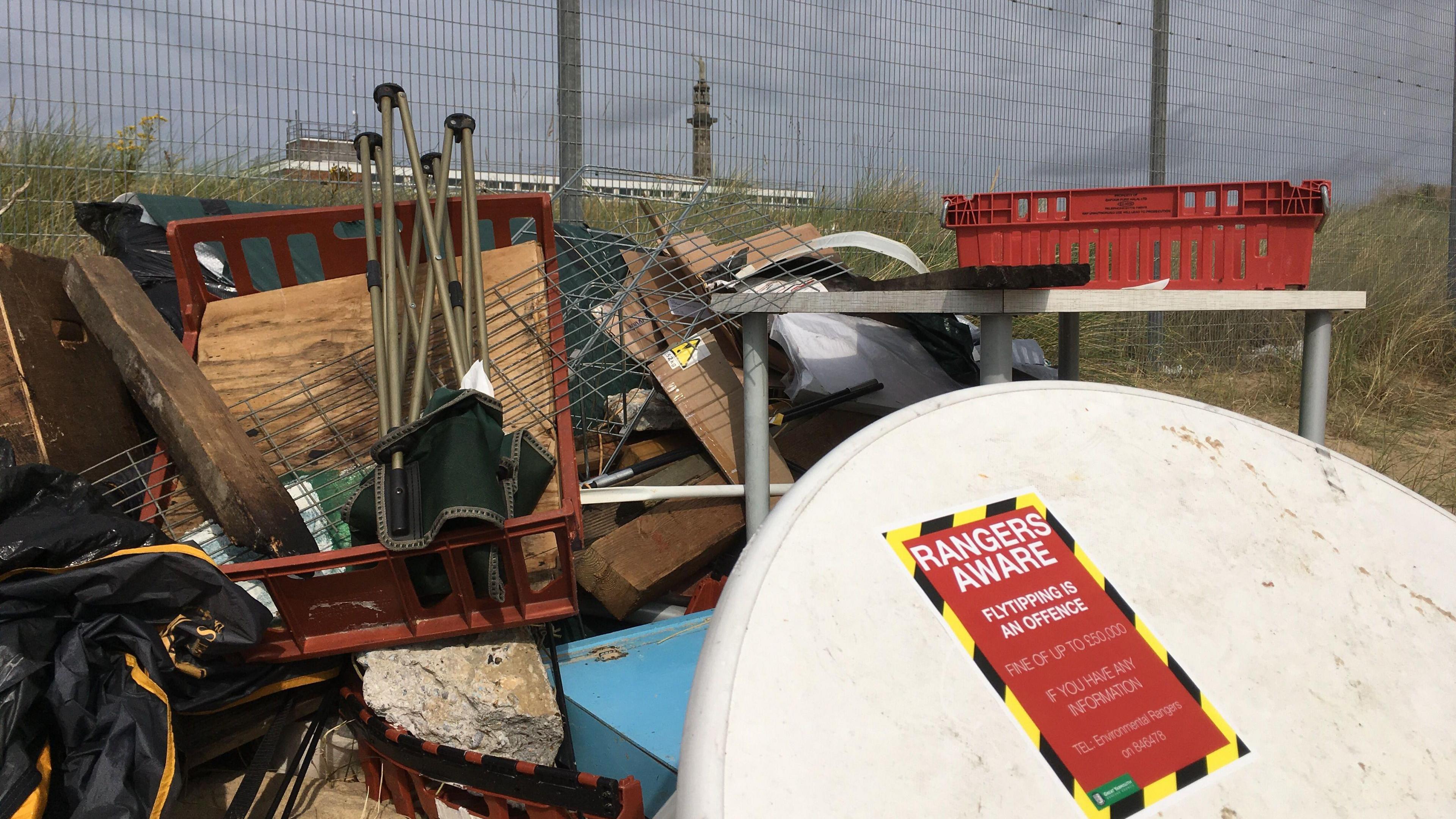 Items including camping chairs and tables are on top of a pile of rubbish fly-tipped on a beach. The table has a red label on it stating that rangers are aware. 