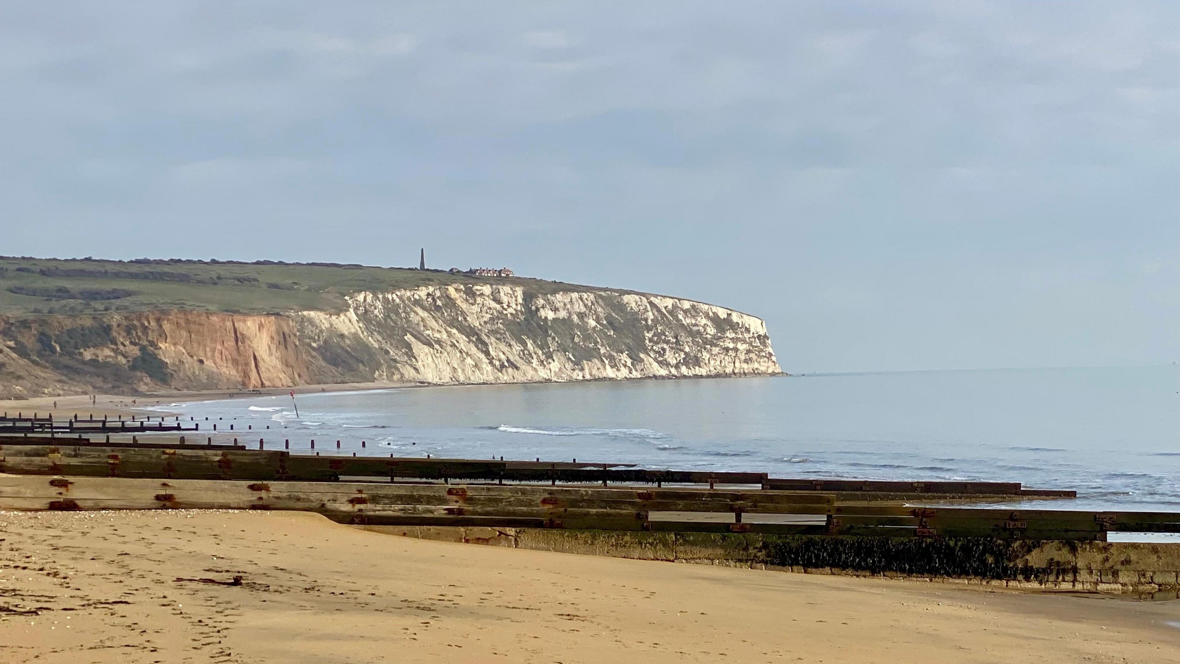 A beach scene on a sunny day. You can see the yellow sand and groynes stretching into the distance. At the end of the beach you can see a white cliff. On top of the cliff is a row of coastguard cottages and the Culver monument. 