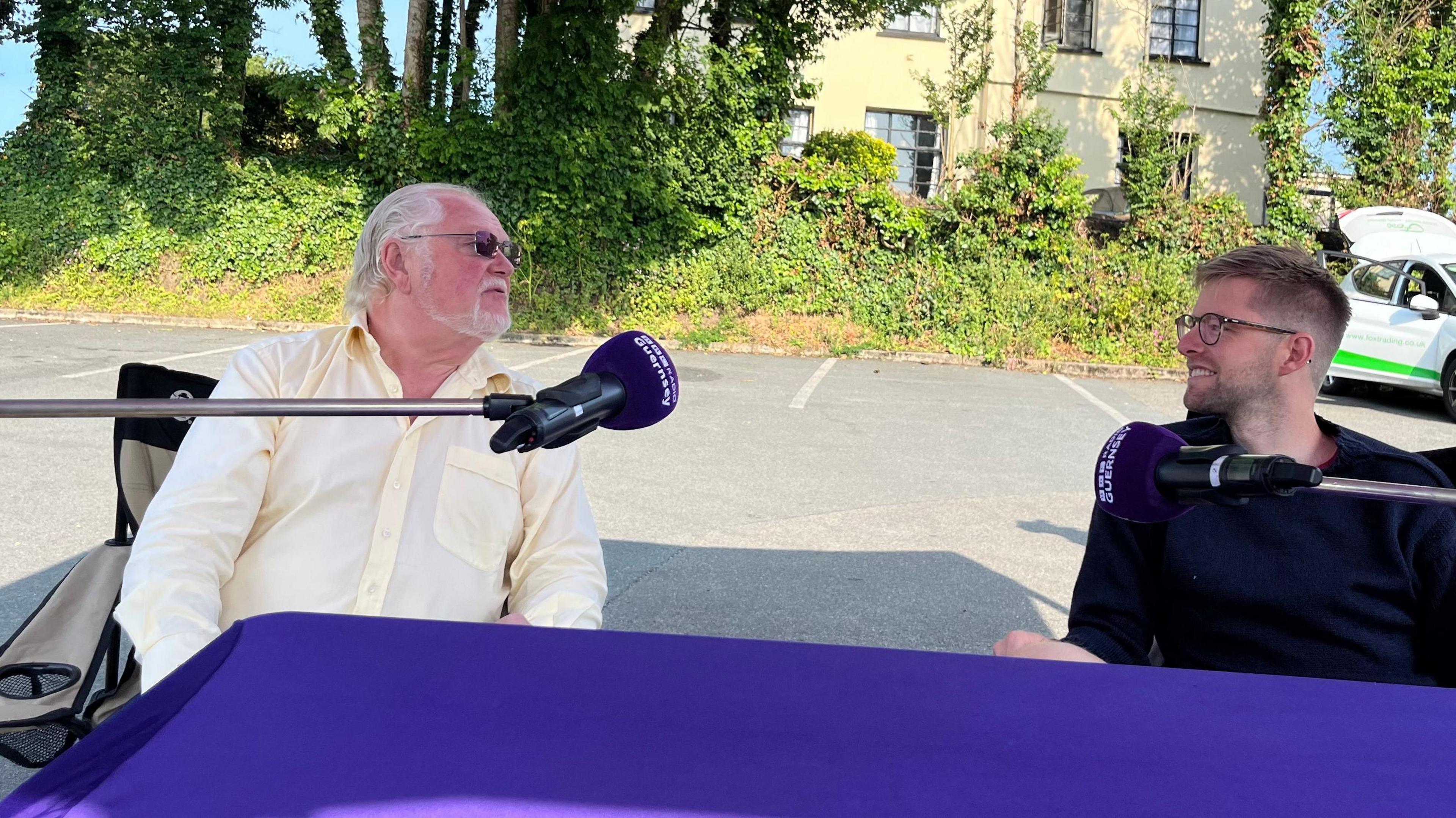 Two men arguing behind a branded purple BBC Radio Guernsey microphone. On the left is Lord Digby Jones, he has white shoulder length hair and a yellow shirt and on the right is Deputy Andy Taylor, he's wearing a blue Guernsey and glasses and has spiky blonde hair and a beard. 