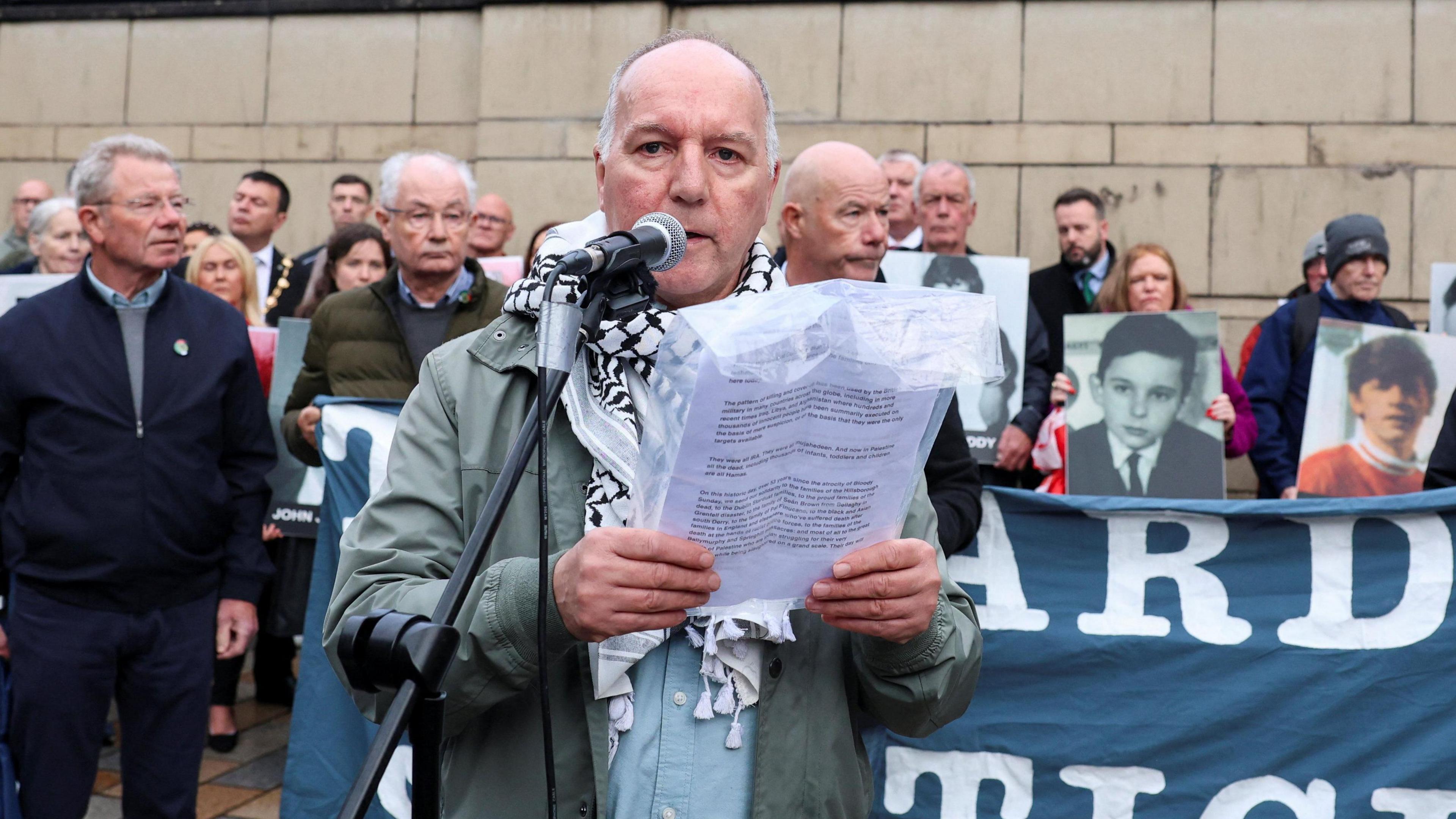 Tony Doherty outside court speaking from a sheet of paper. He has short white/grey hair and is wearing a green jacket and blue top with a keffiyah scarf. (It is black and white)