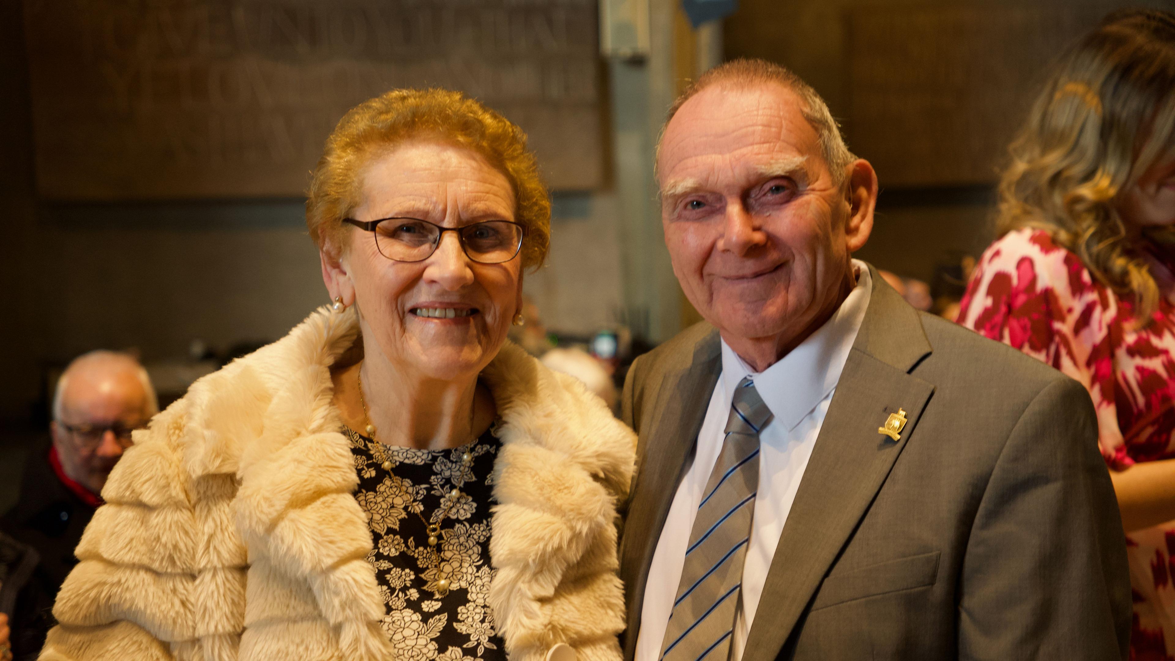 A woman in a white coat and black dress, with glasses, and a man in a grey suit, white shirt and grey tie with thin blue stripes