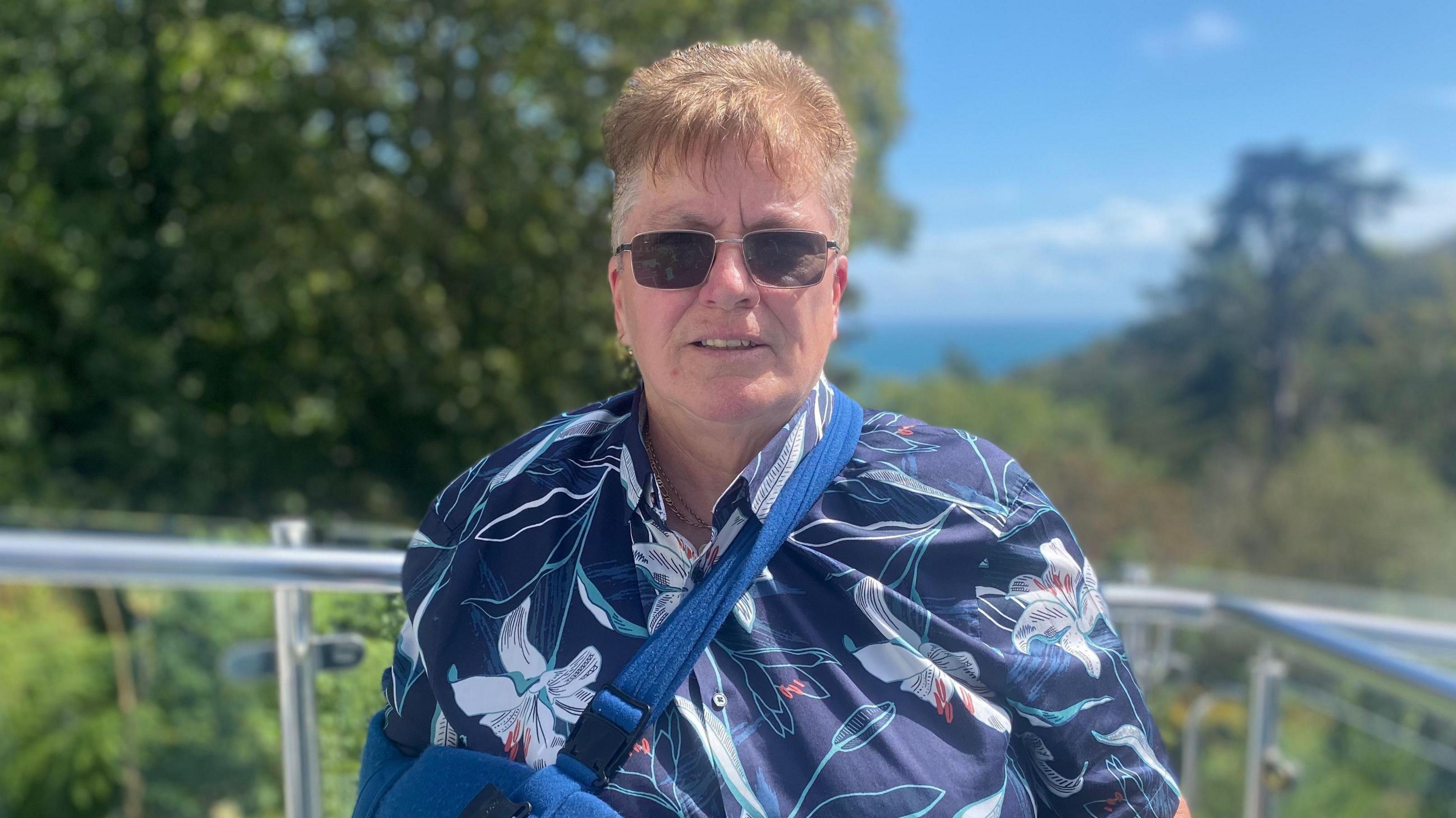 Linda Garnham stands on a balcony in front of several trees and other plants. She has sunglasses on and a blue floral shirt. Her right arm is in a blue sling. It is a sunny day.
