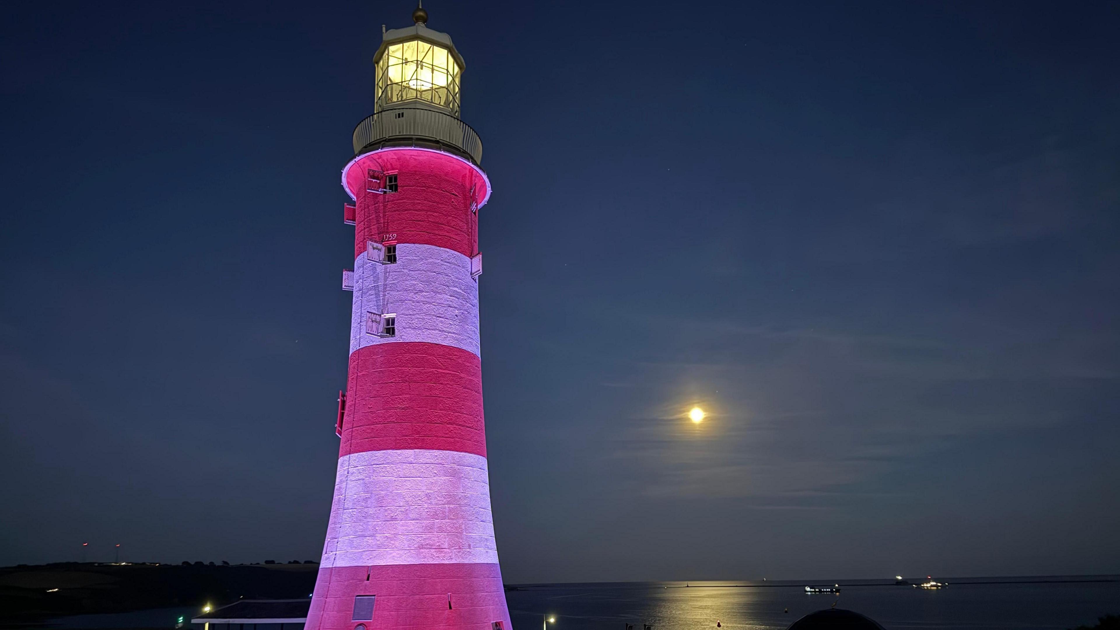 Plymouth lighthouse lit up at night. In the background is the sea, with the moon reflecting on the water. The lighthouse is red and white and there is a light on at the top.