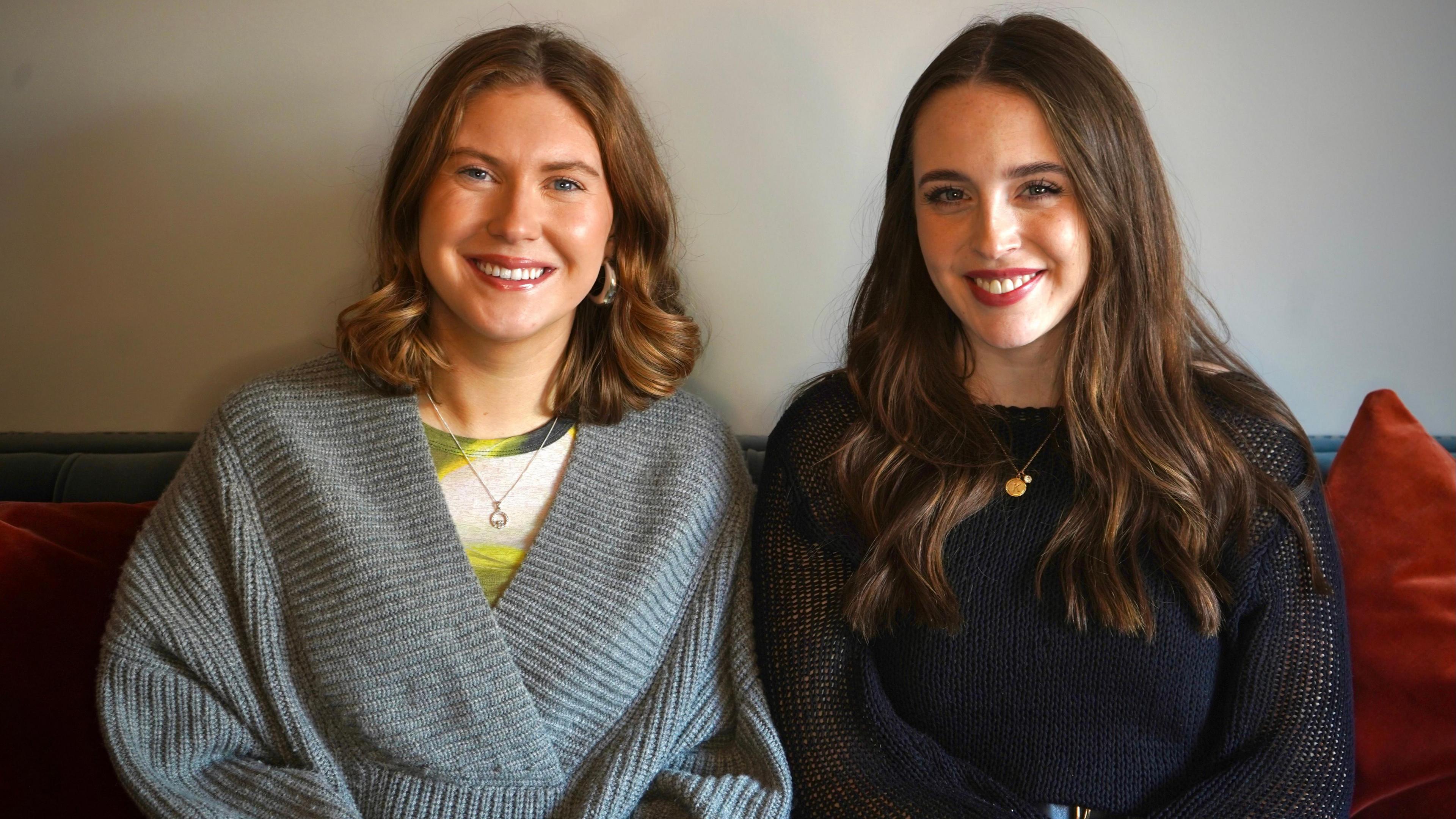 Two women sit facing the camera. Both are smiling. One woman is wearing a grey jumper while the other woman is wearing a black jumper.