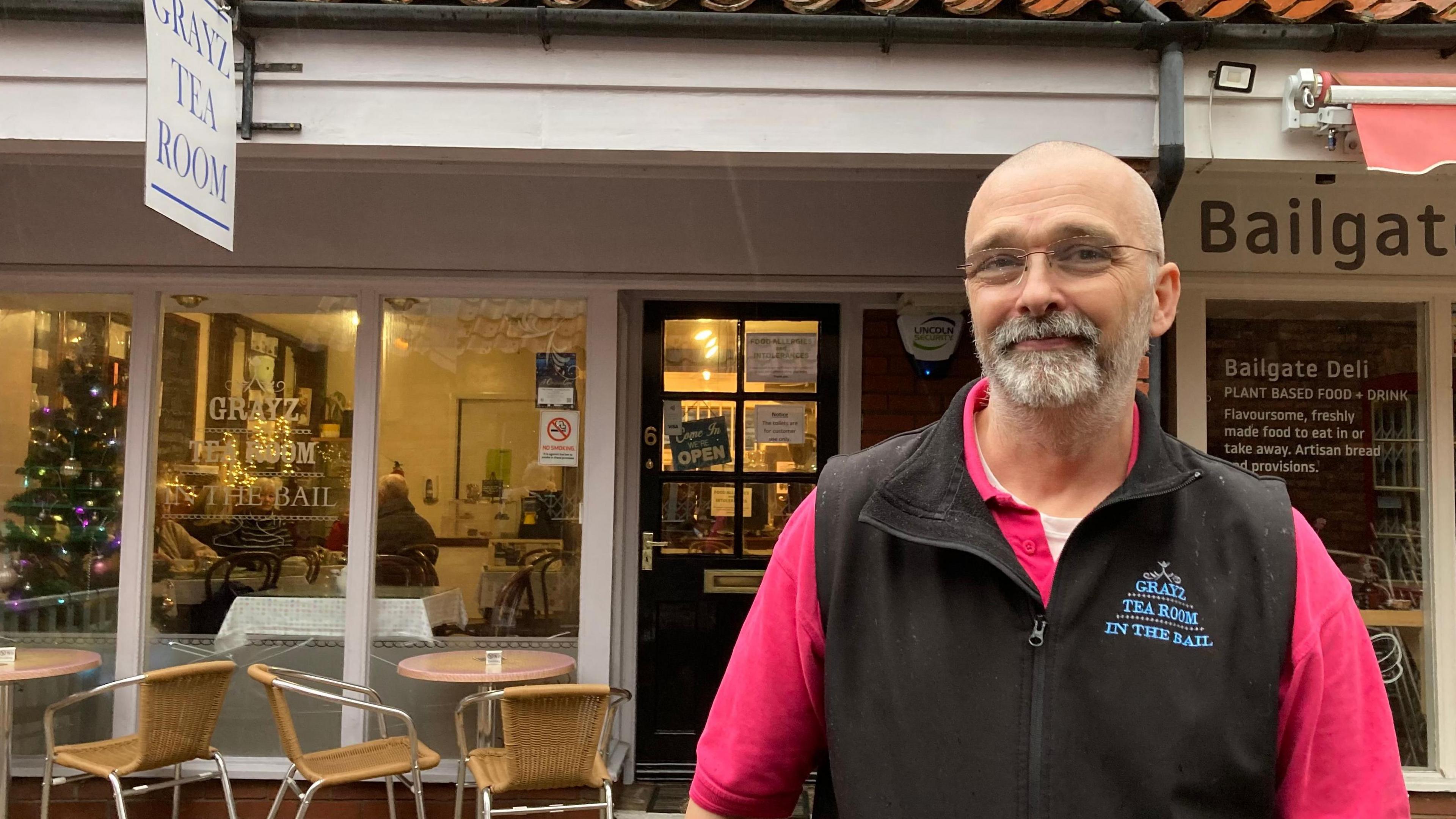 Scott Eversfield, owner of Grayz Tea Room, standing outside the business wearing a black gilet with the company name printed on it. He has a goatee-style beard and glasses