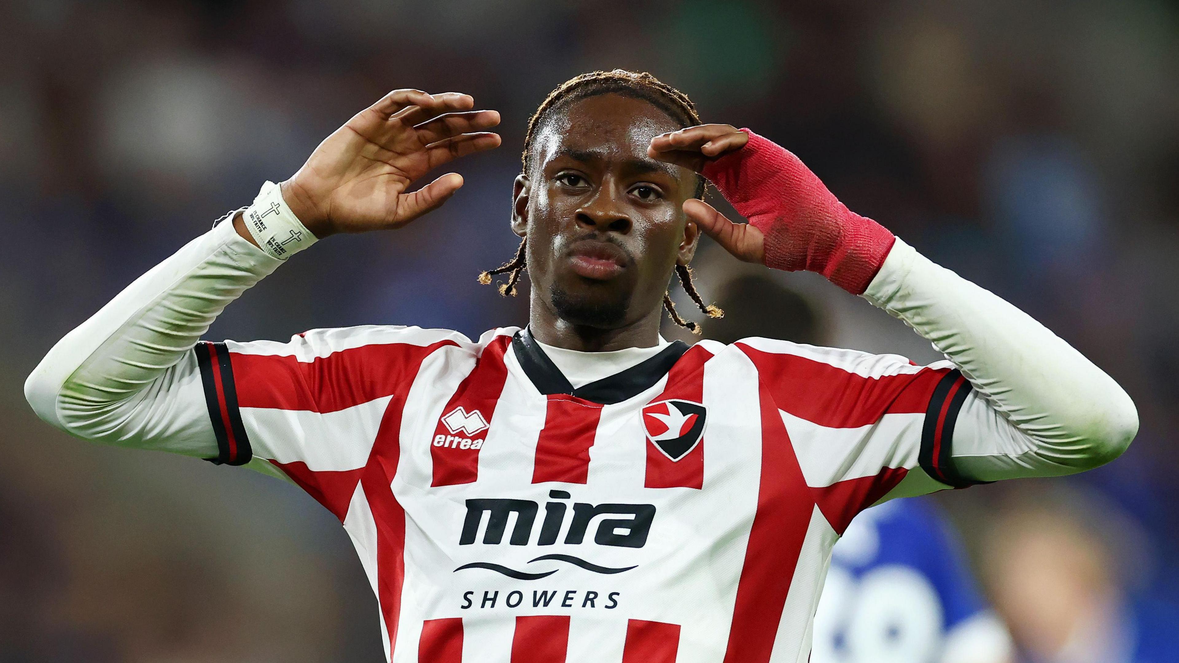 A Cheltenham Town player, wearing the club's red and white striped top, holds his arms above his head during a match away at Cardiff City