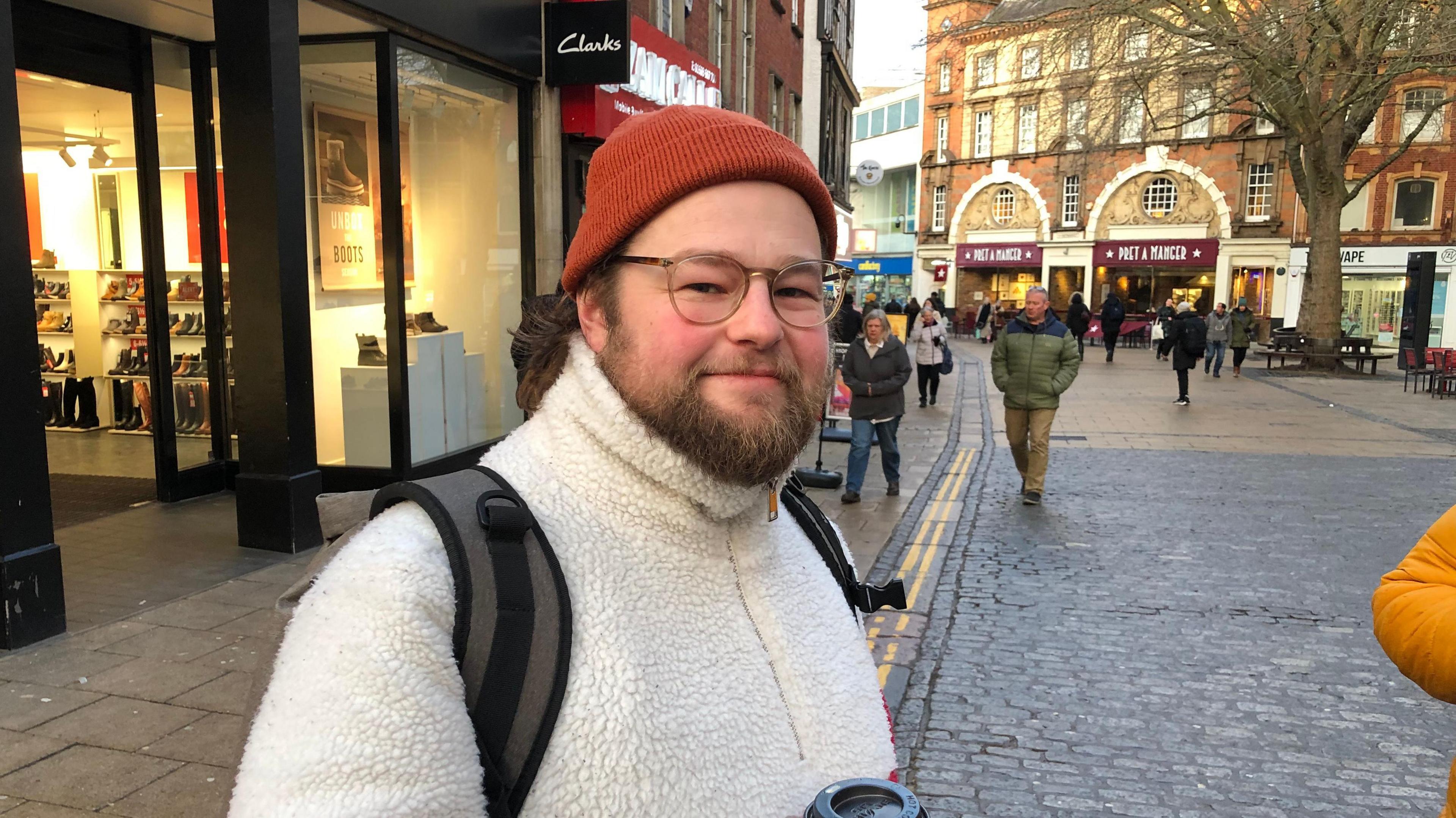 Adam is is Norwich city centre. He's wearing a reddish-brown beanie hat, glasses and a white fleece jacket with a backpack.