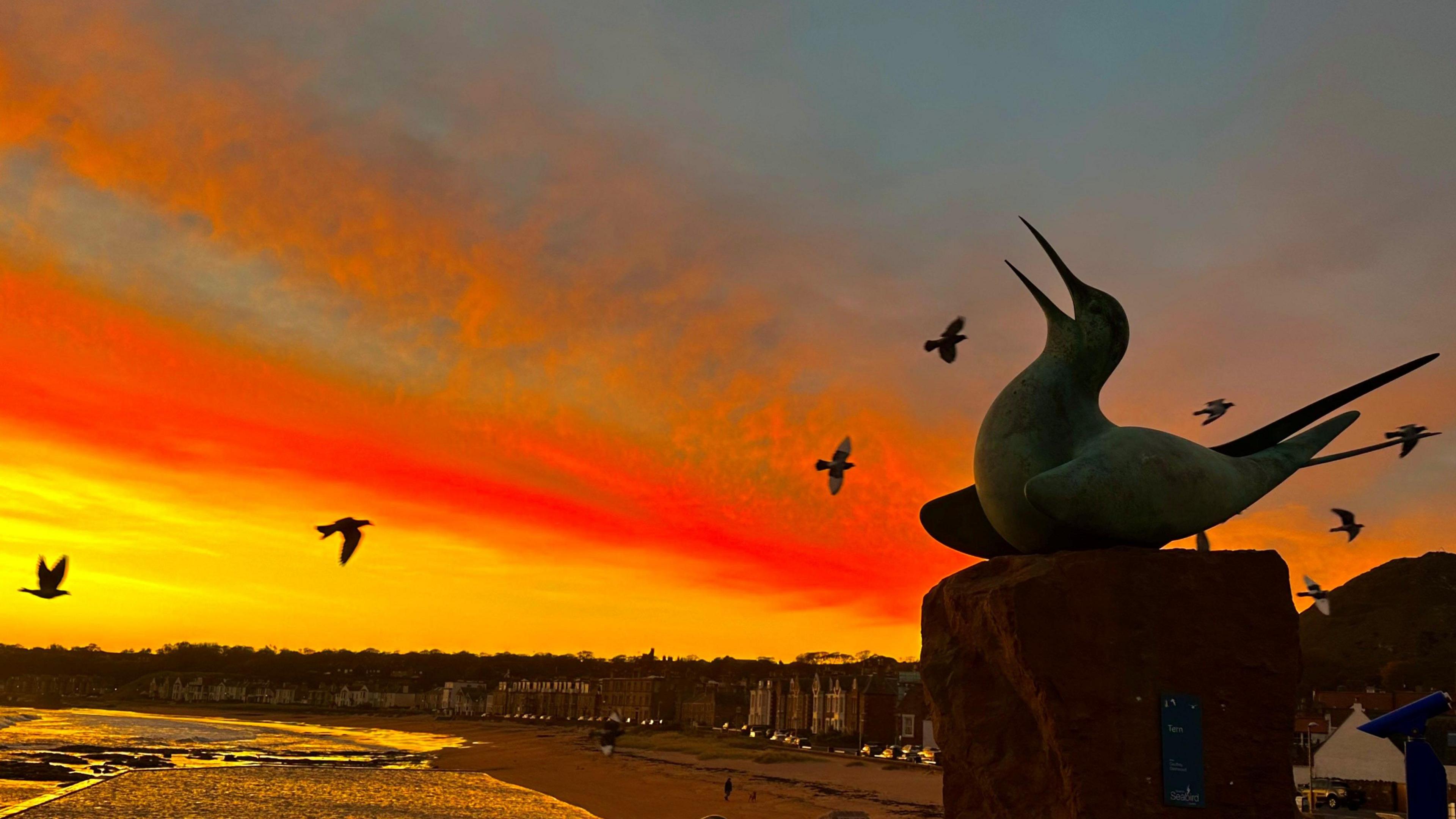 A bird statue is in silhouette against a red and yellow sky. There is water below.