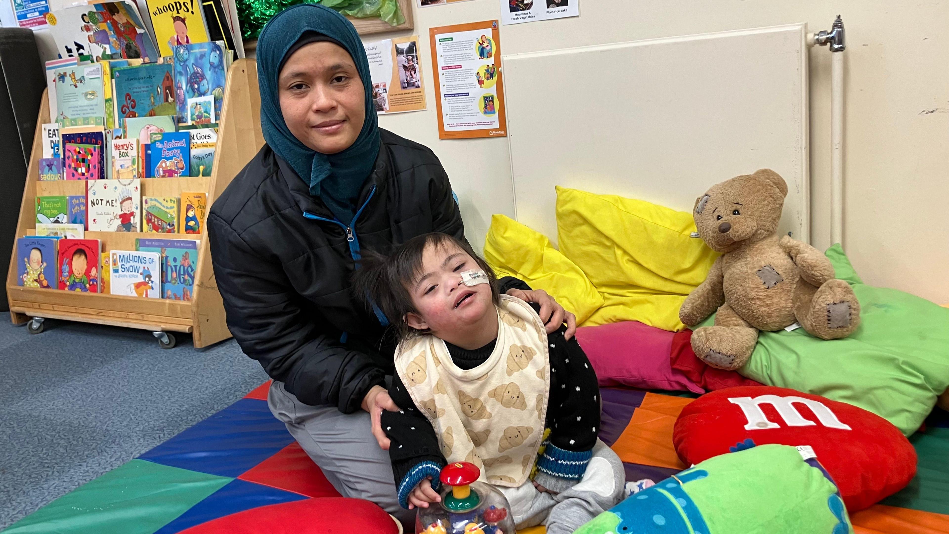 Leni wearing a blue head-scarf, a black puffer coat and grey trousers- sitting on a colourful play mat holding her daughter, who is wearing a black cardigan and a yellow bib.