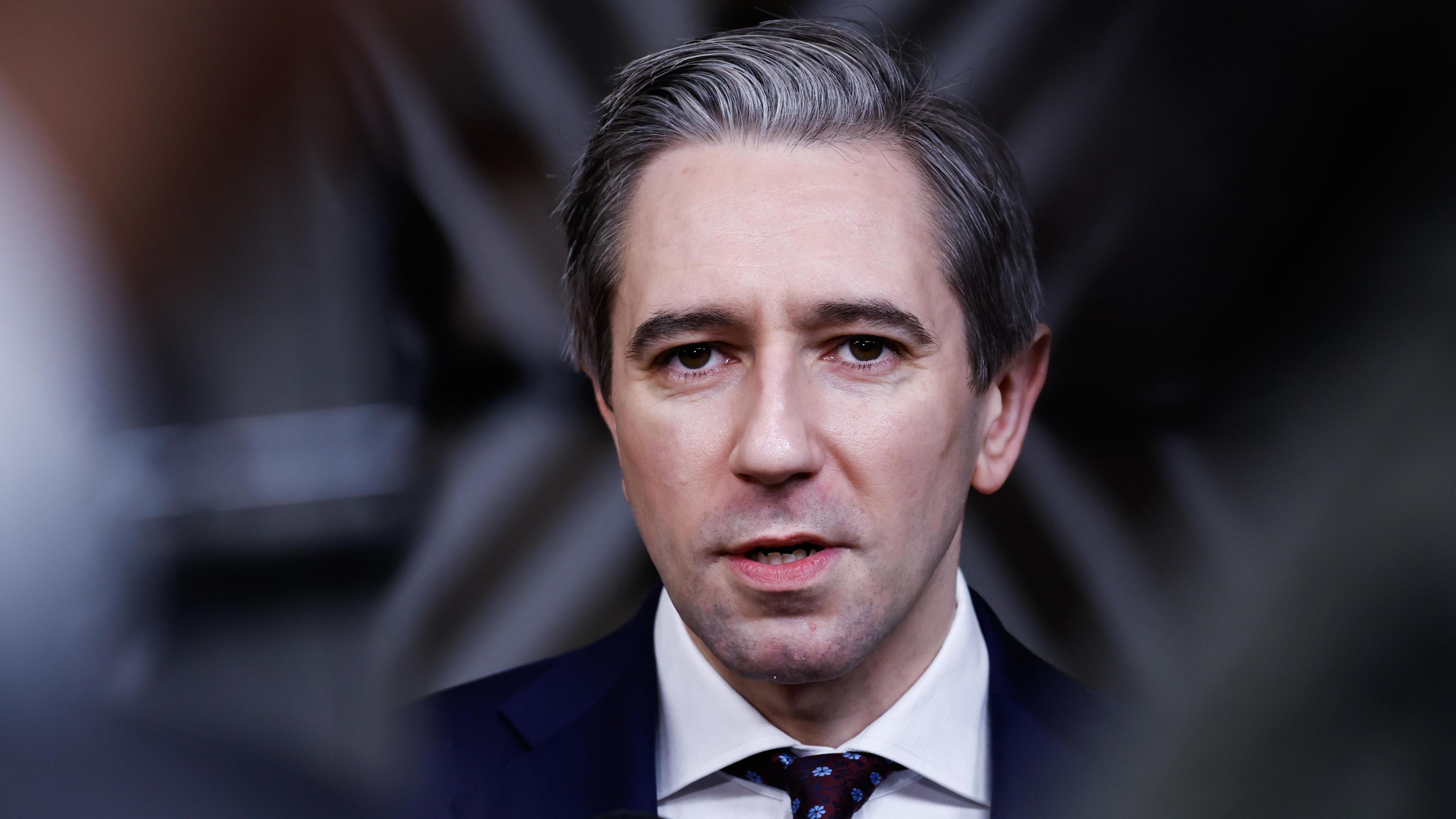 Simon Harris - a man with short, grey hair. He is mid-speak and is looking slightly beyond the camera. He is wearing a dark suit jacket, a patterned dark tie and a white collared shirt.