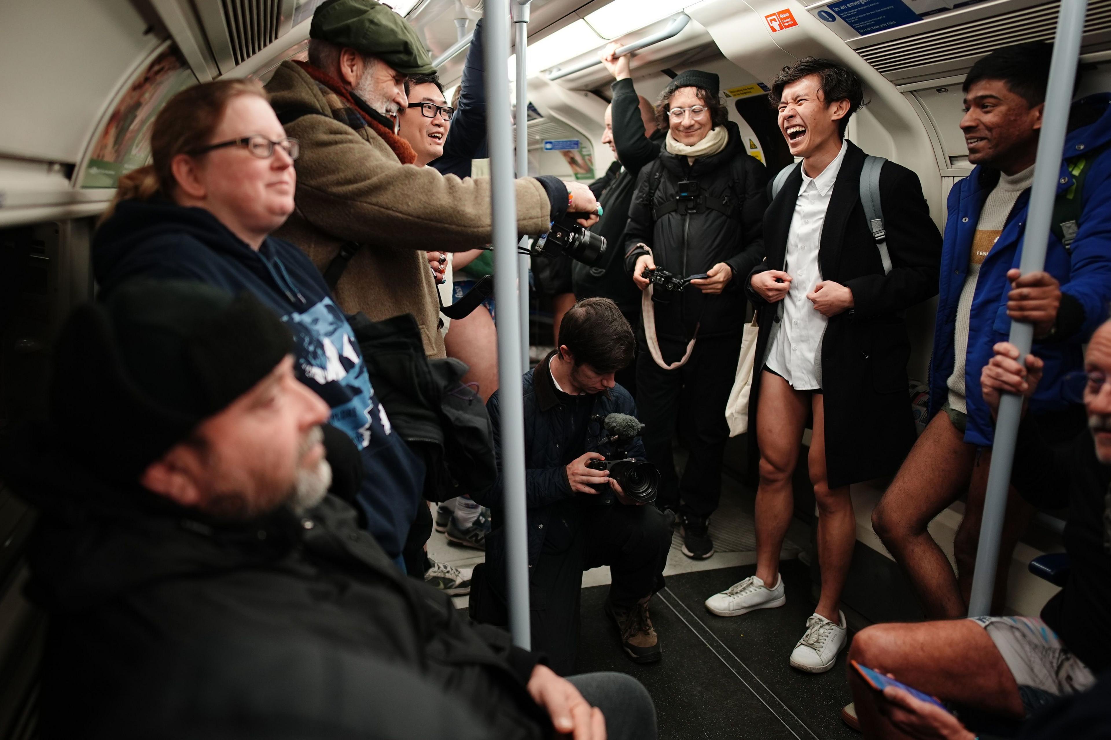 Two men stand on the Tube with no trousers (right), while in the middle photographers take pictures and to the left a seated man and standing woman look away