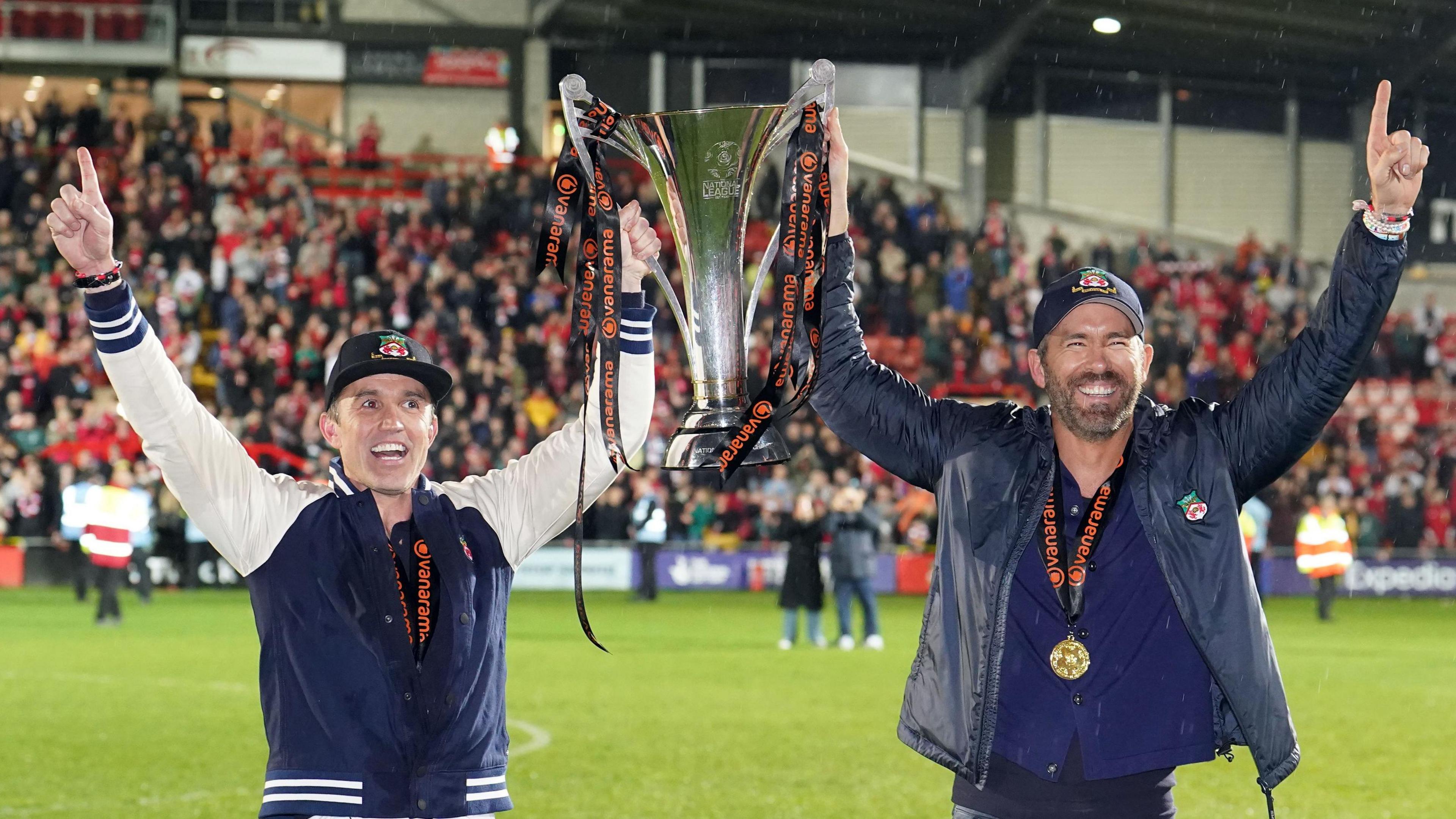 Picture of two men, Hollywood stars Rob McElhenney and Ryan Reynolds, dressed in sports jackets and Wrexham football club branded baseball caps holding aloft a silver cup on a football pitch against the background of football fans in the stand behind them. 