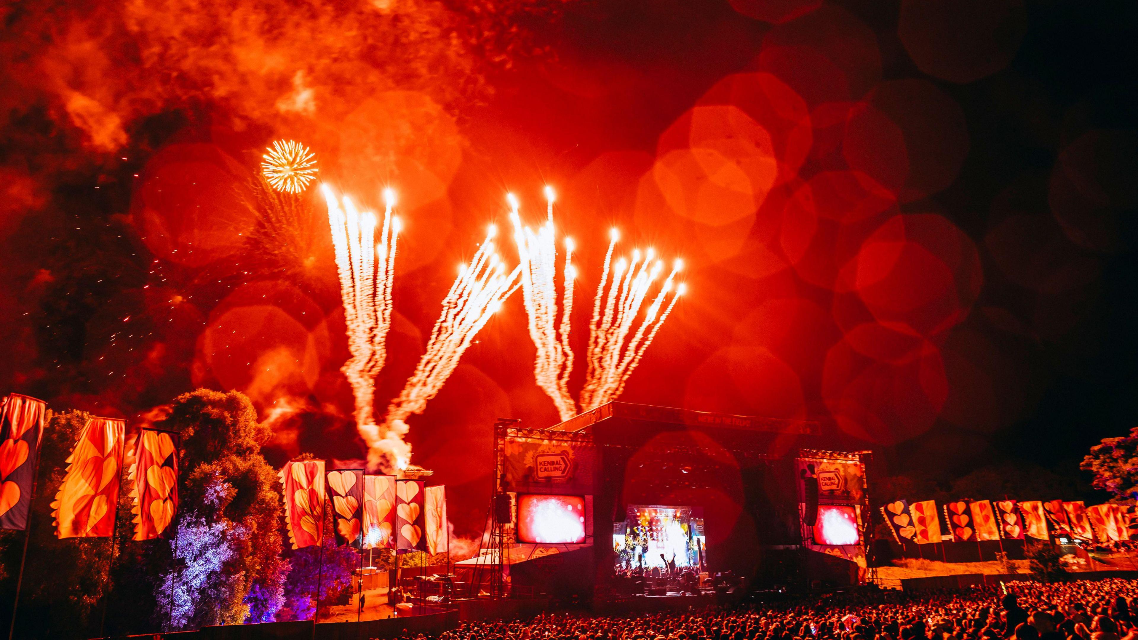 A Kendal Calling Main Stage with huge crowds in front. It is night time and red fireworks light up the night sky.