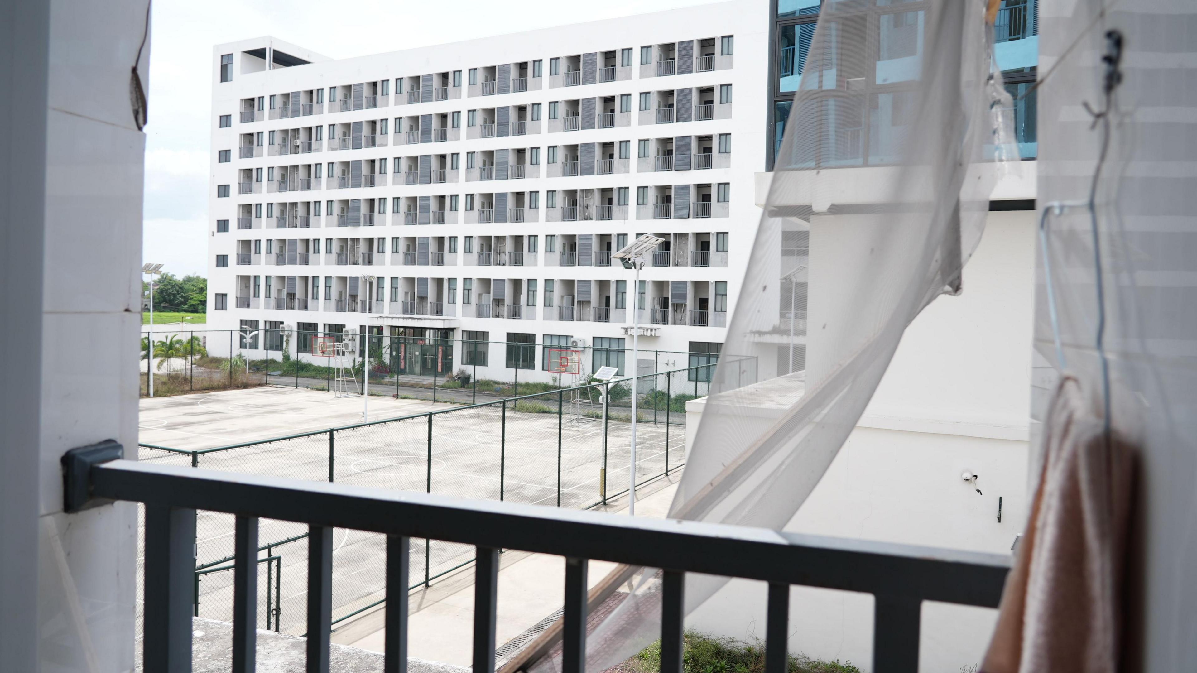 A view from a balcony, with a ripped curtain and an old towel in the foreground. Beyond these can be seen a basketball court, behind which lies another dormitory block several floors high