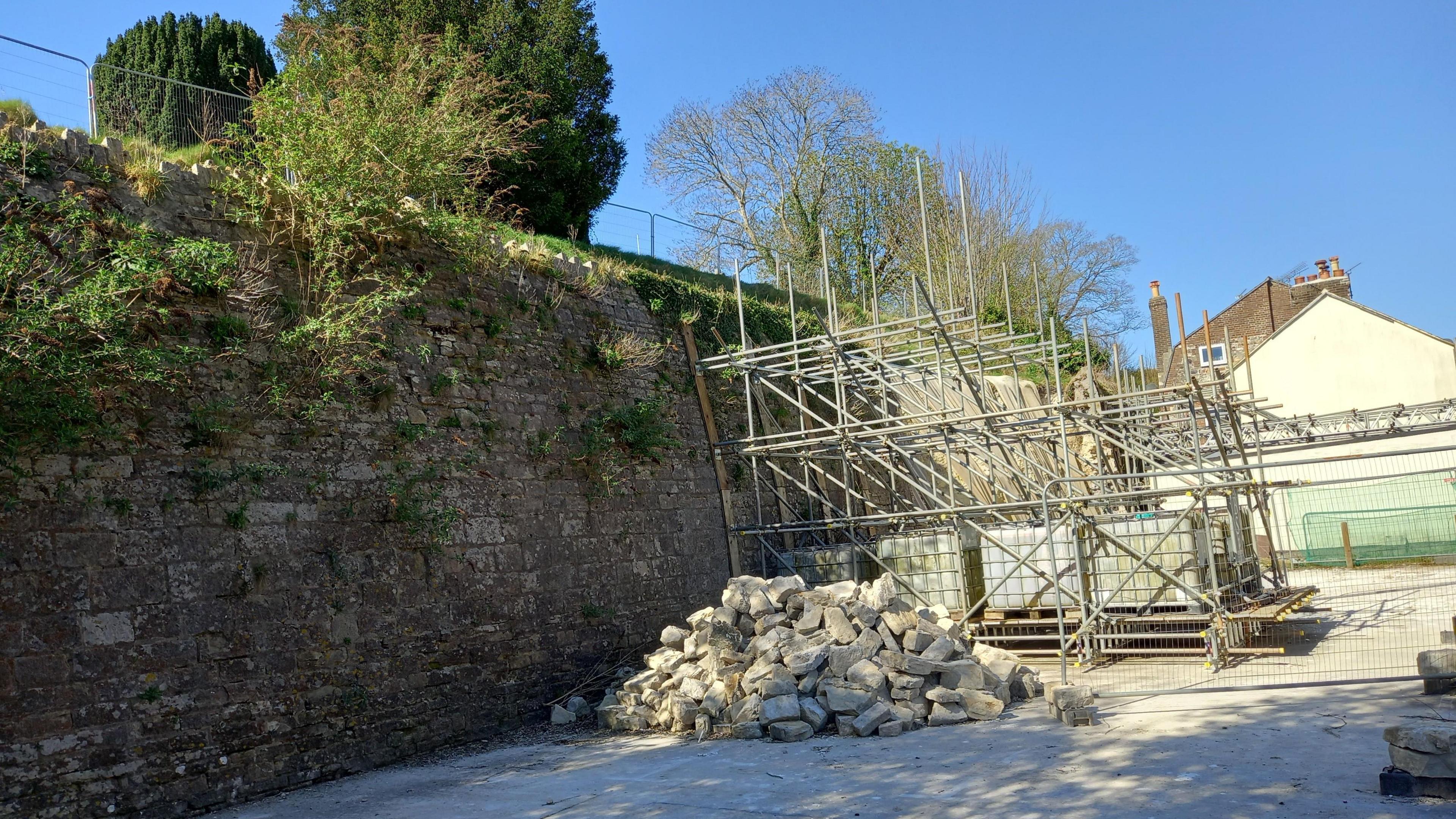 A view of the Fordington Cemetery with the supporting scaffolding in Holloway Road. A pile of rubble lies next to the scaffolding. It is sunny.

