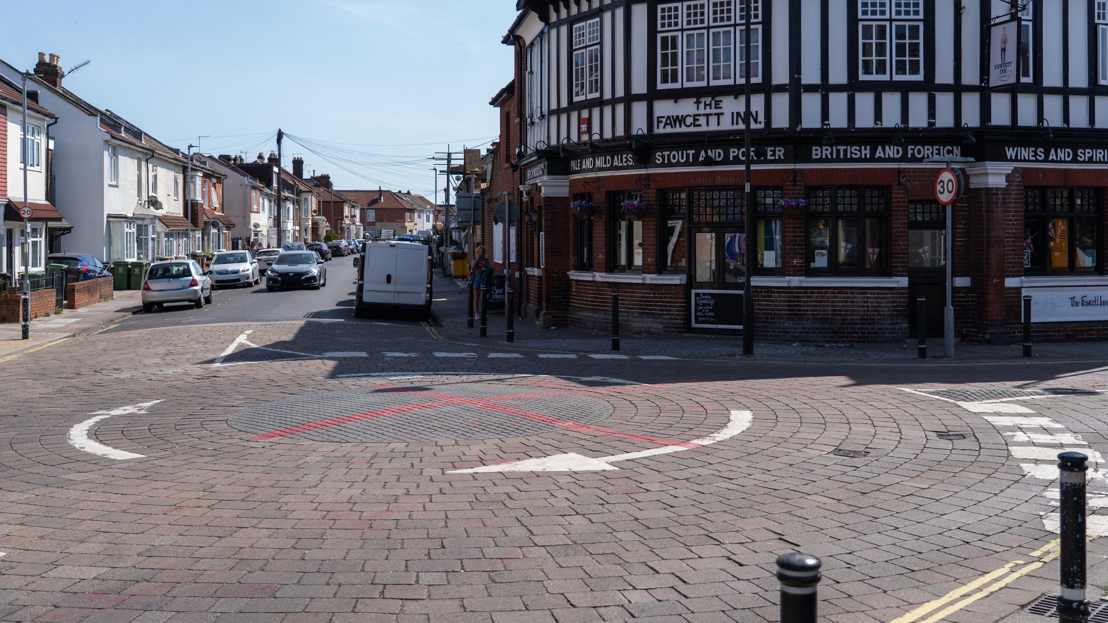 A black roundabout with white arrows painted around it on a cobble street in Portsmouth. The central circle of the roundabout has red graffiti paint on it in a cross. On the edge of the roundabout is The Fawcett Inn Pub and a residential street in the distance.