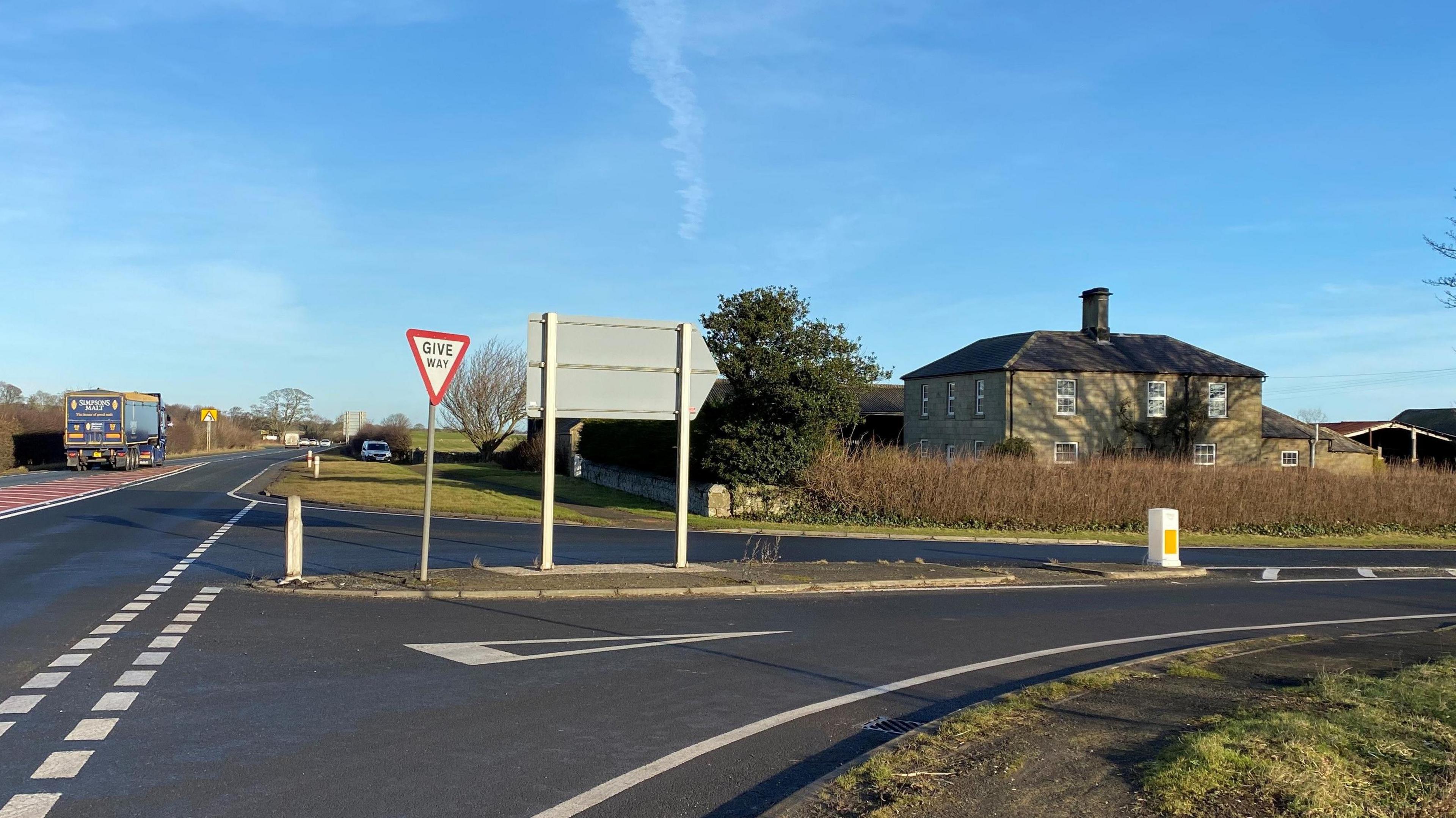 A very large square two-storey Georgian farmhouse set back from the A1 behind a hedge sits on a busy junction with a lorry passing.