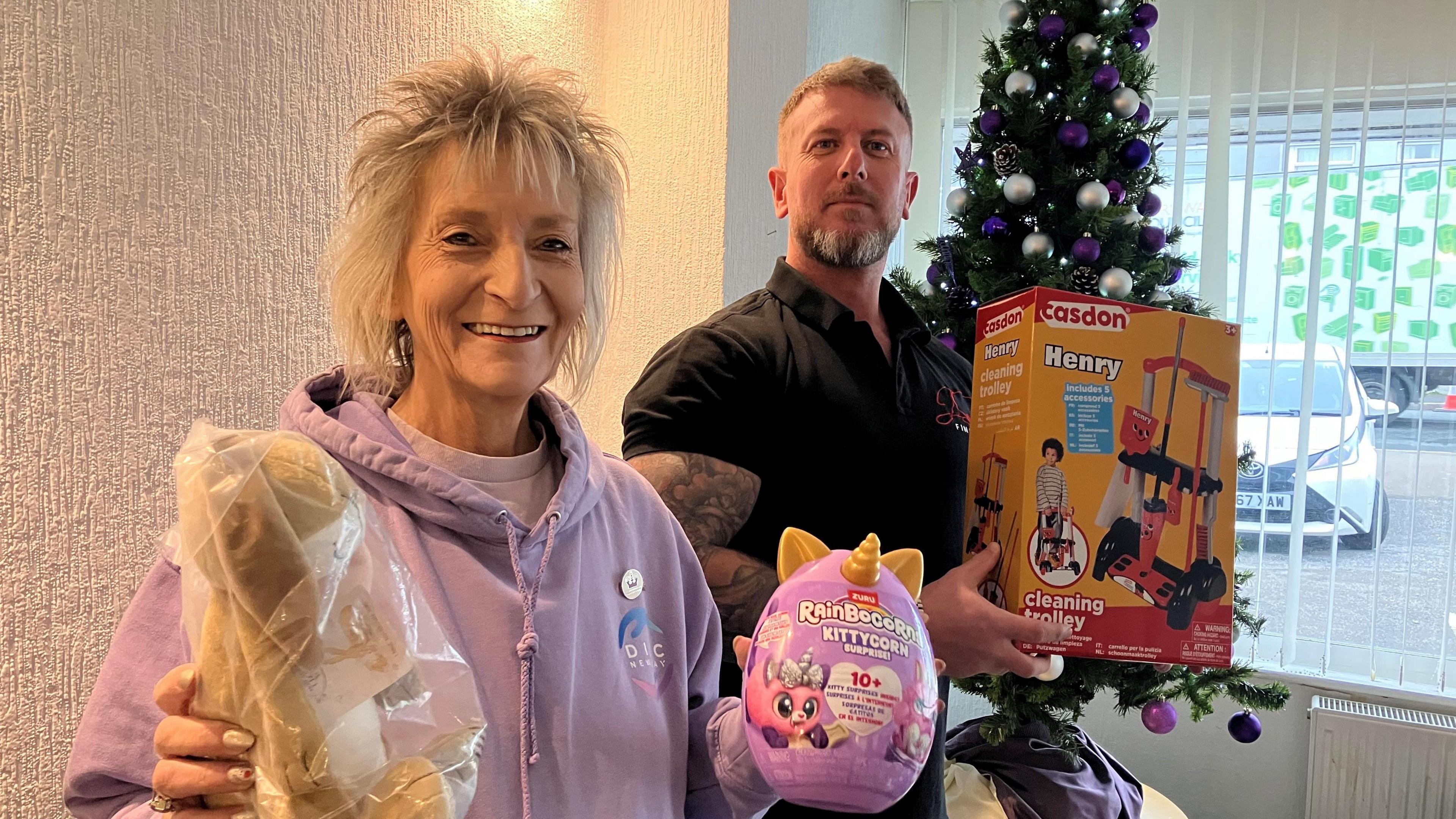 Woman holding a soft toy in a bag and a plastic Rainbocorns toy and a man holding a child's cleaning trolley 